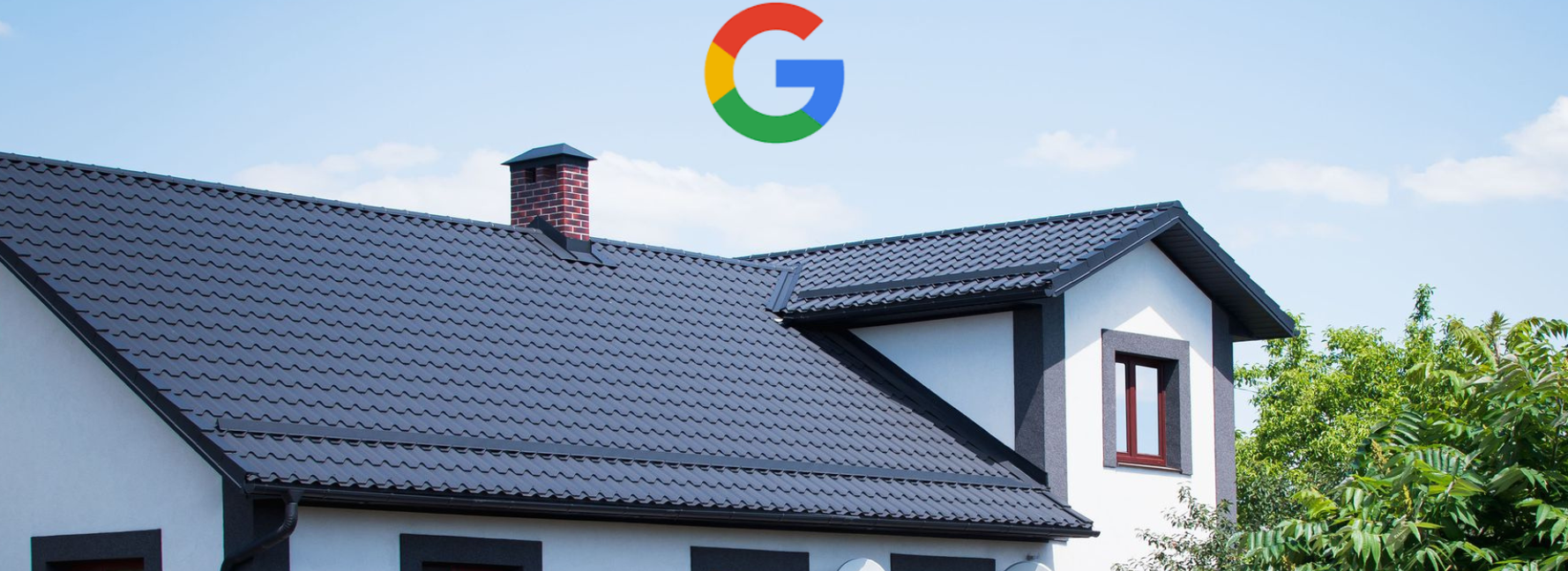 A white house with a dark, textured roof and a chimney against a blue sky, partially obscured by green trees.