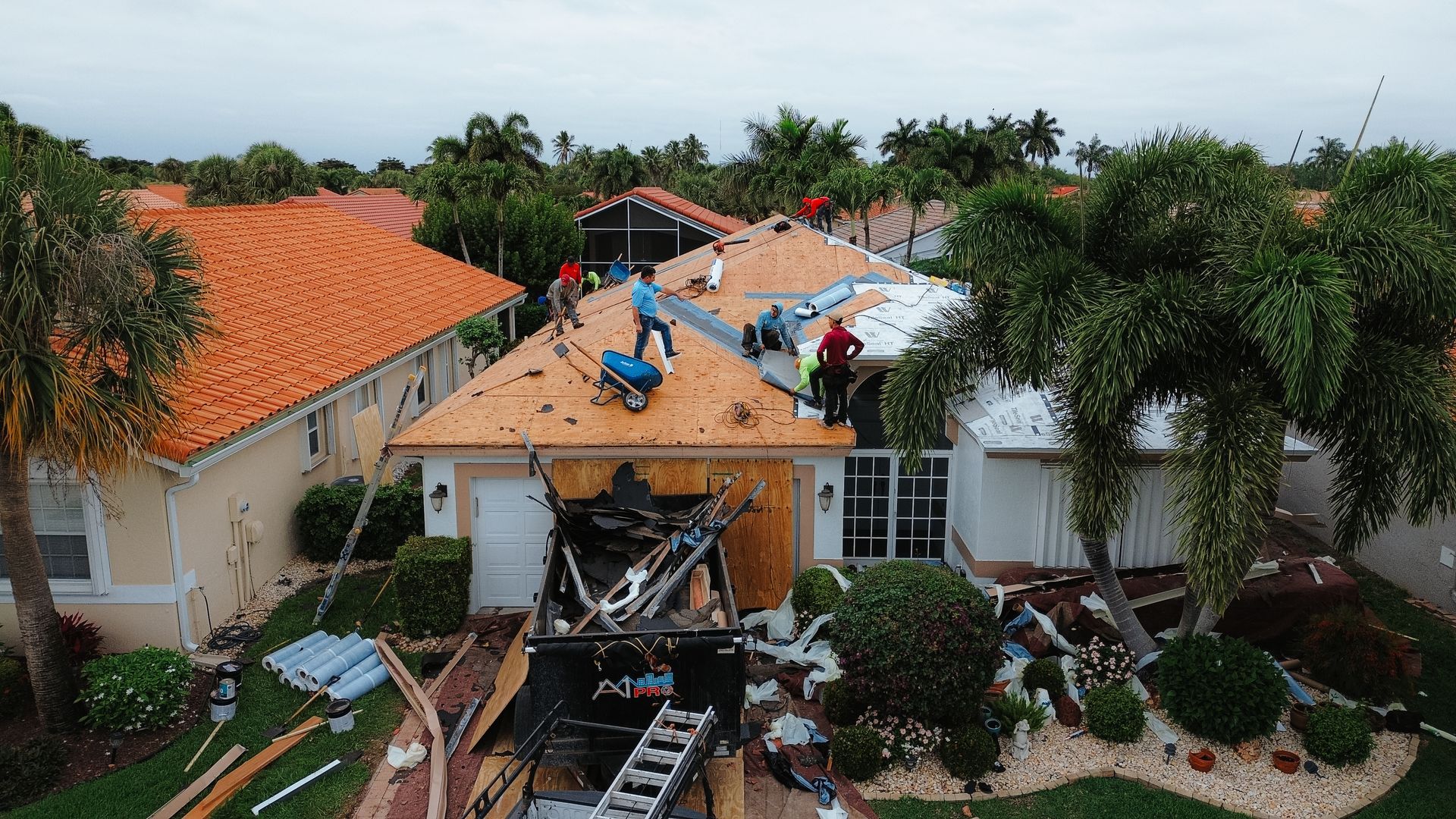 A high-angle view of a gray asphalt shingle roof on a sunny day, with a glimpse of a nearby house and backyard pool below.