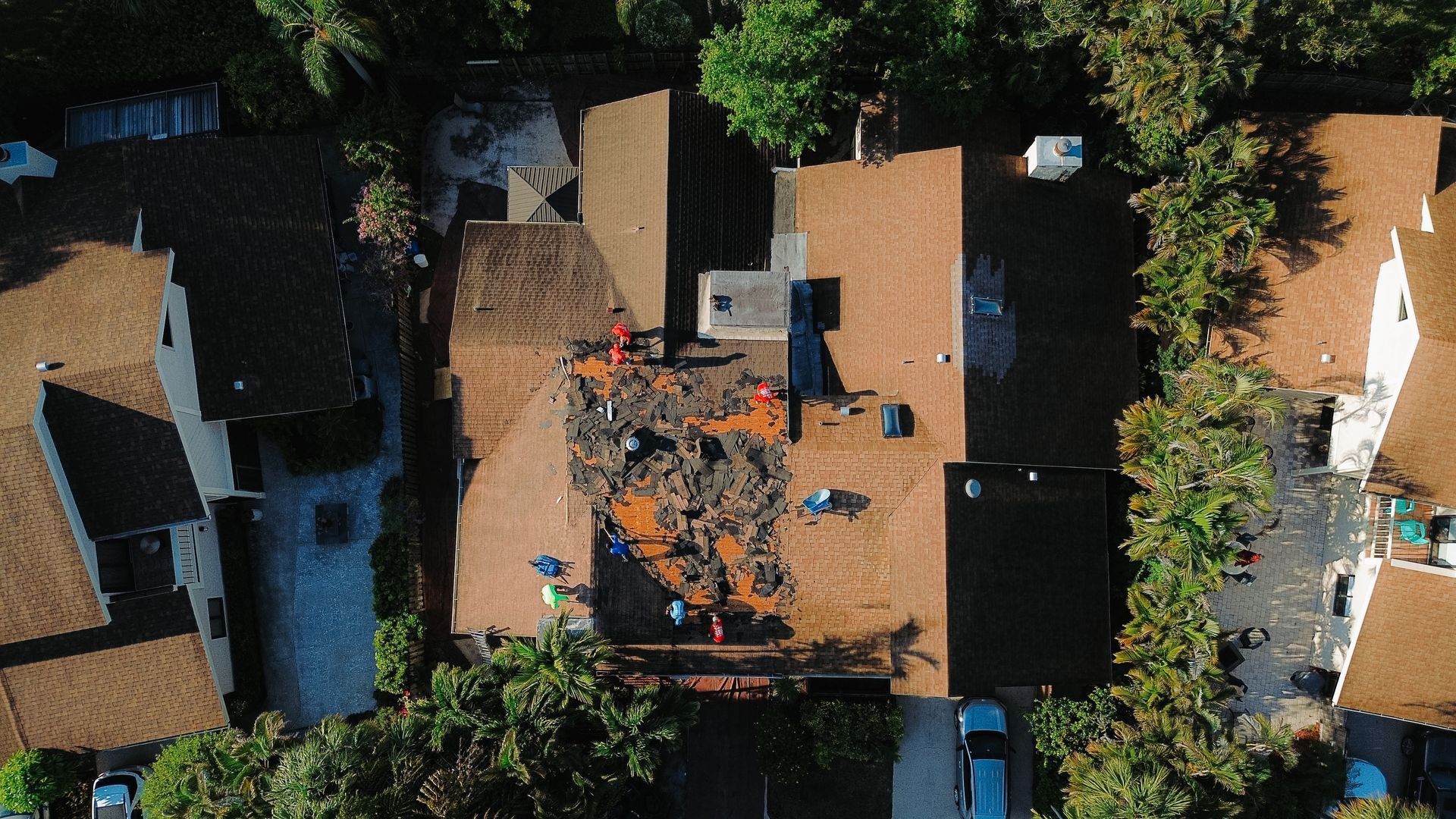 A person in a blue shirt applies sealant around a plumbing vent pipe on a flat, dark roof with many circular fasteners.