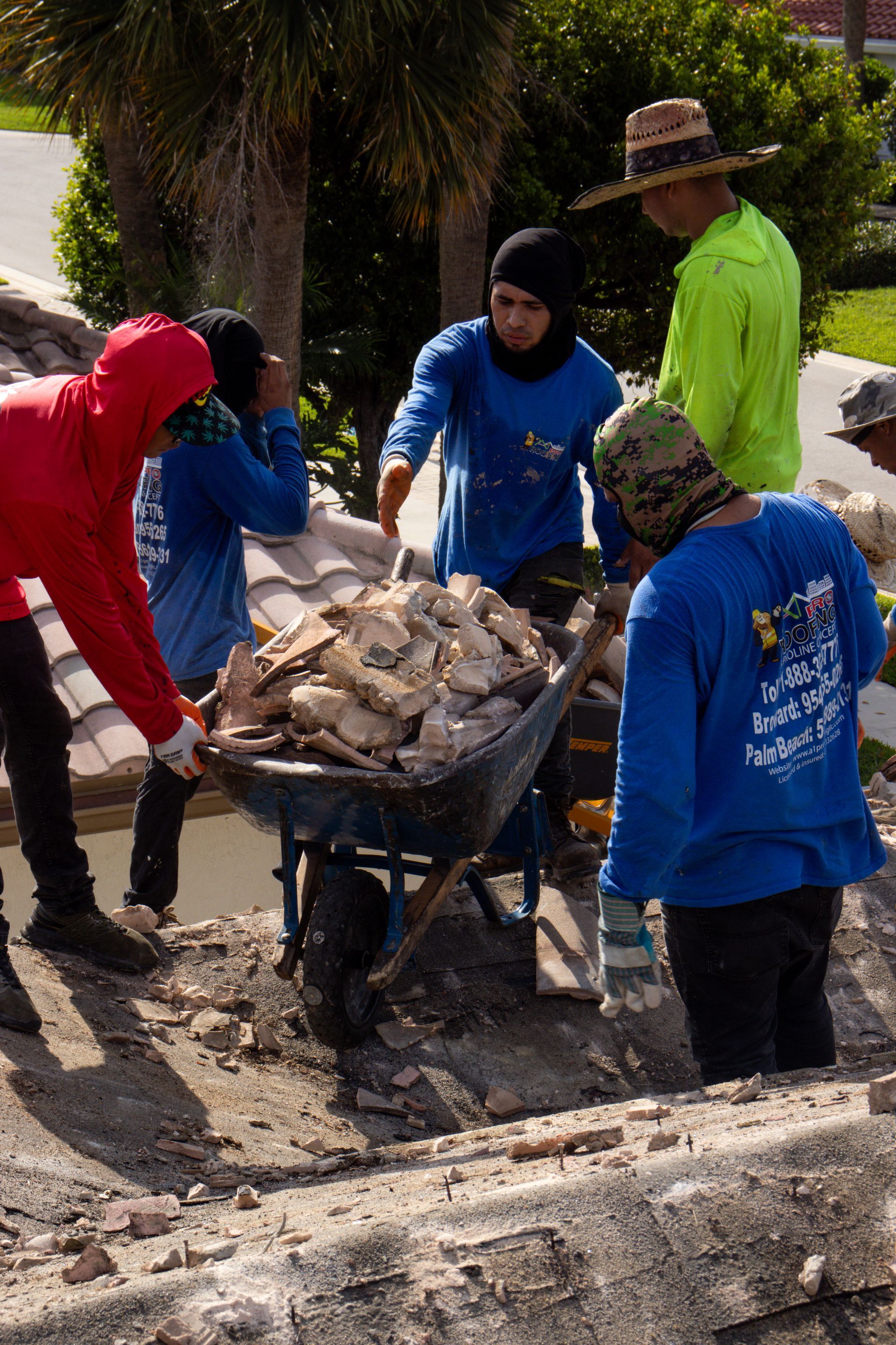 A person in a blue shirt applies sealant around a plumbing vent pipe on a flat, dark roof with many circular fasteners.