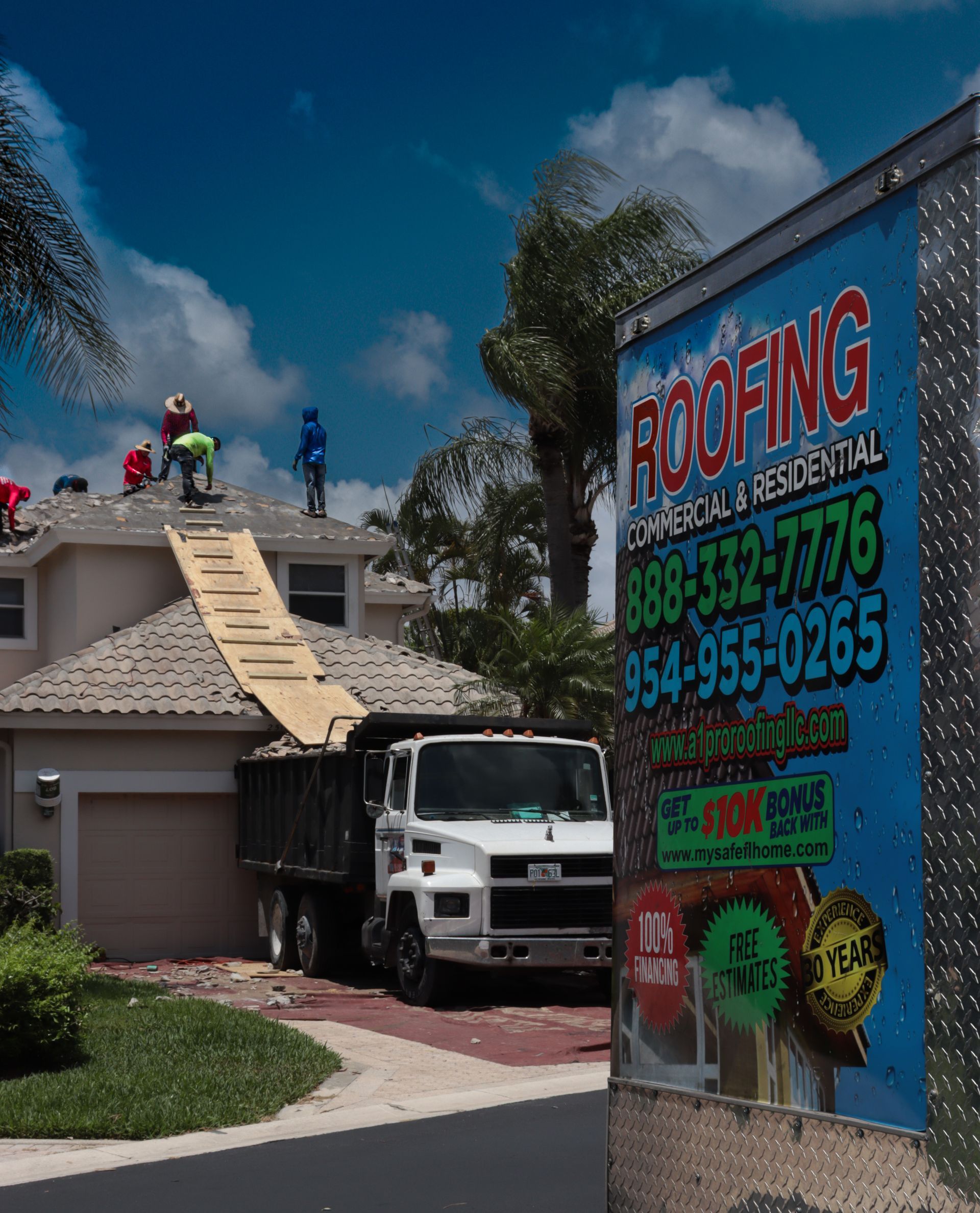 A worker in a blue shirt and hat uses a shovel to remove old roof shingles into a chute on a sunny, residential roof.
