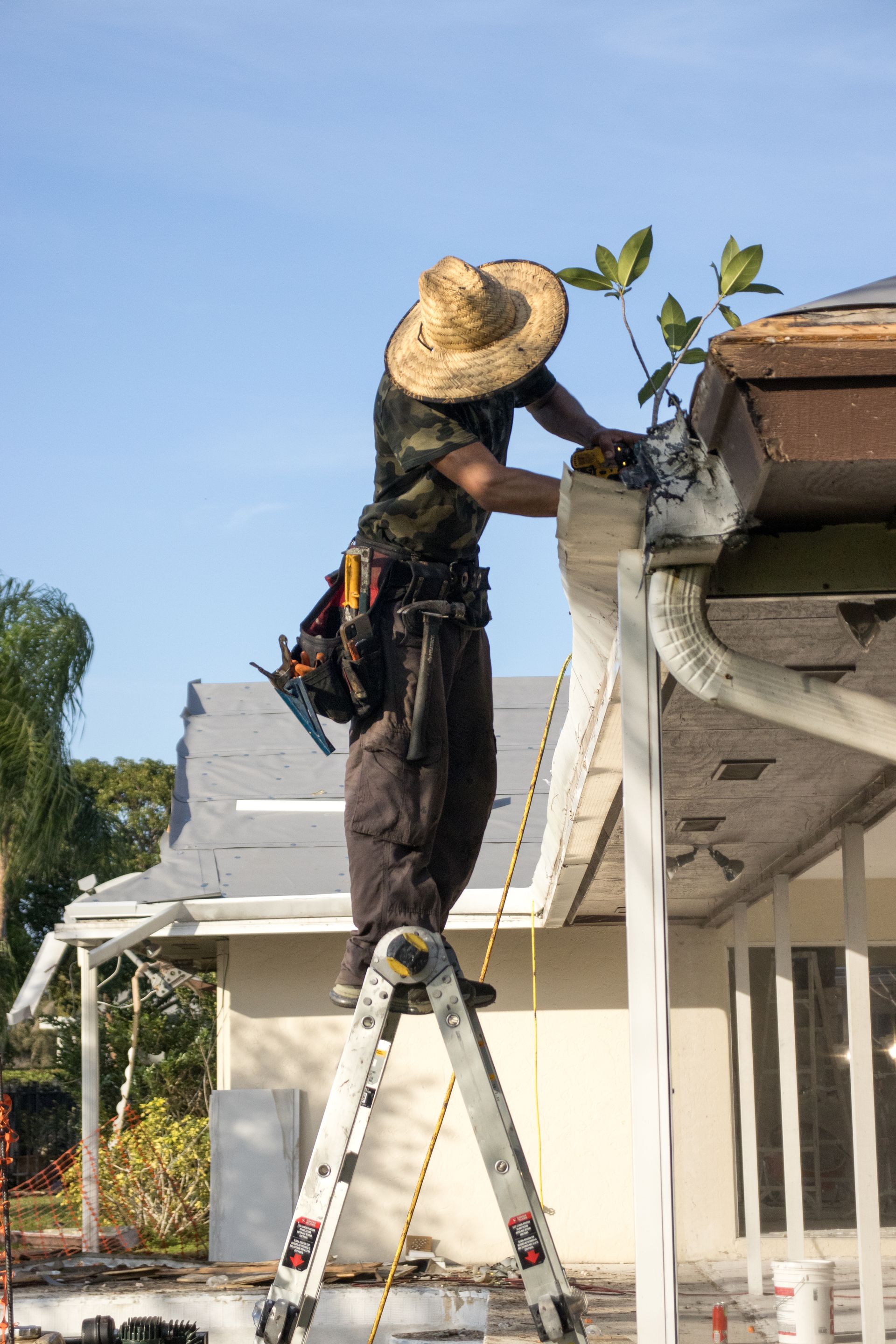 A person on a ladder wearing a grey sweater and dark pants inspecting or repairing the black gutter of a red brick house.