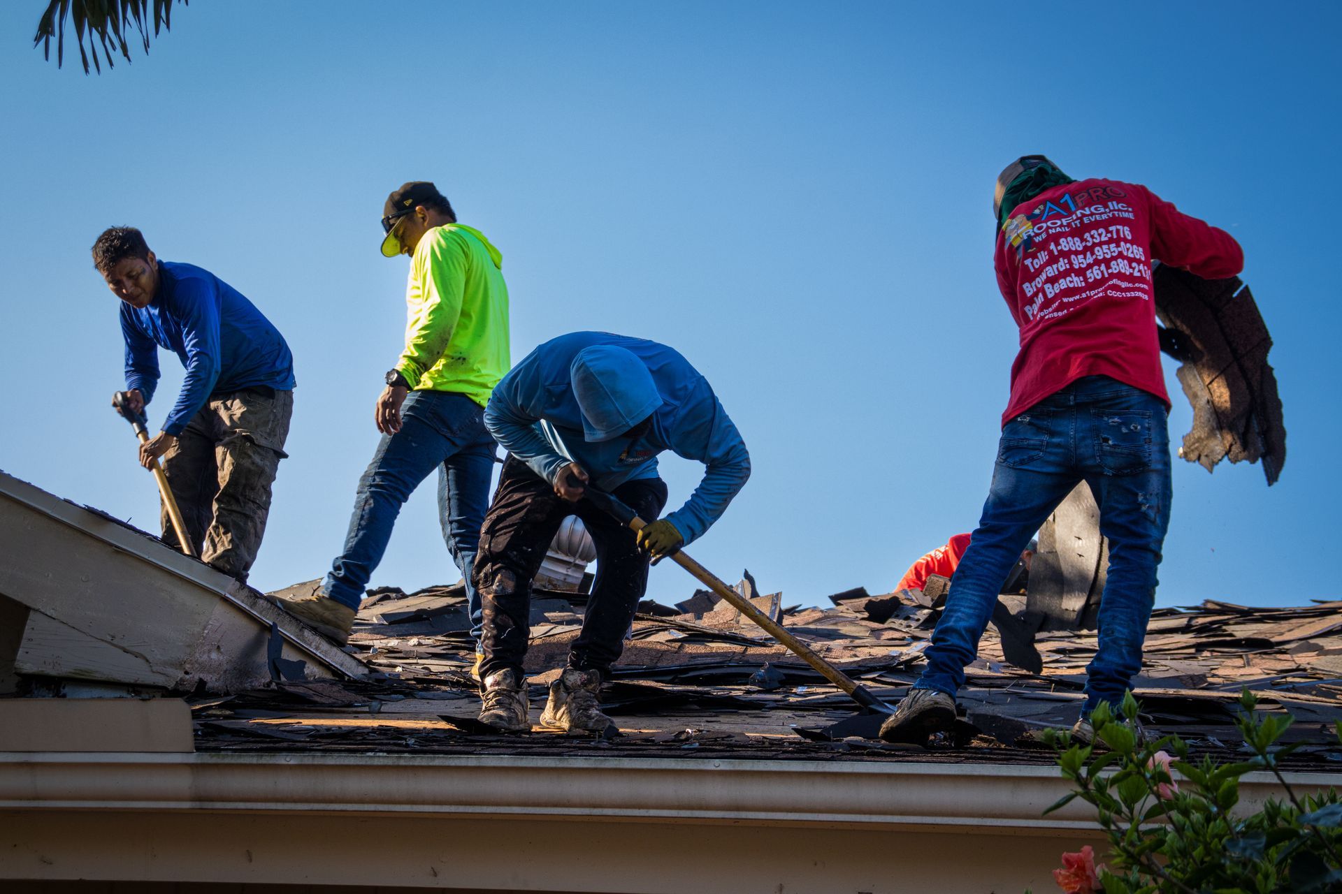 A worker in a blue shirt and hat uses a shovel to remove old roof shingles into a chute on a sunny, residential roof.