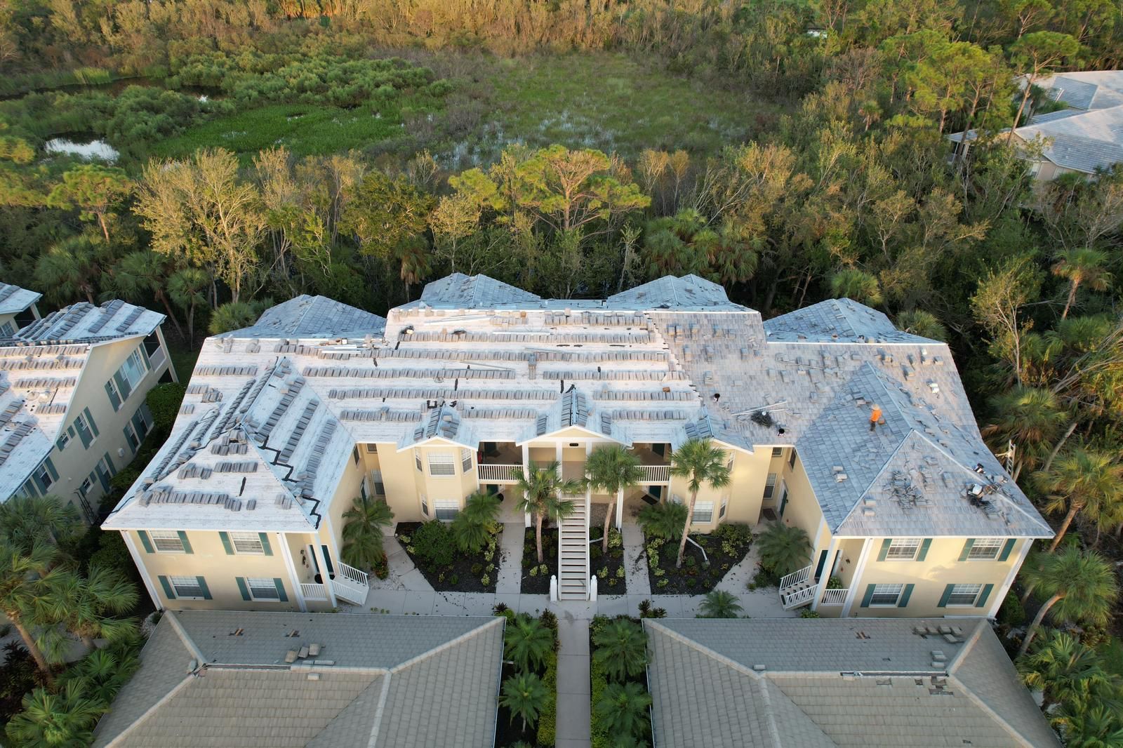 Two roofers in blue hoodies and tan work pants installing red clay tiles on a house roof near a pond.