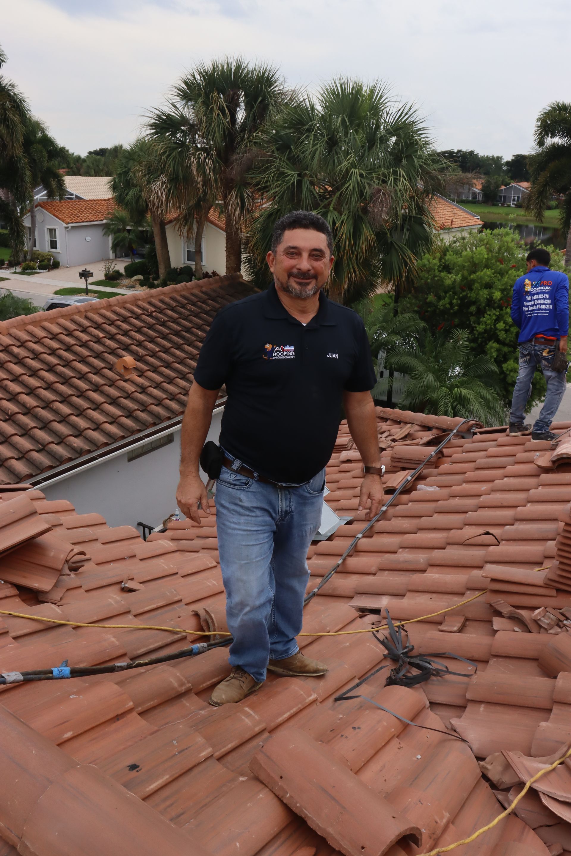 A person in work clothes standing on a wooden roof structure, organizing stacks of grey tiles against a blue sky.
