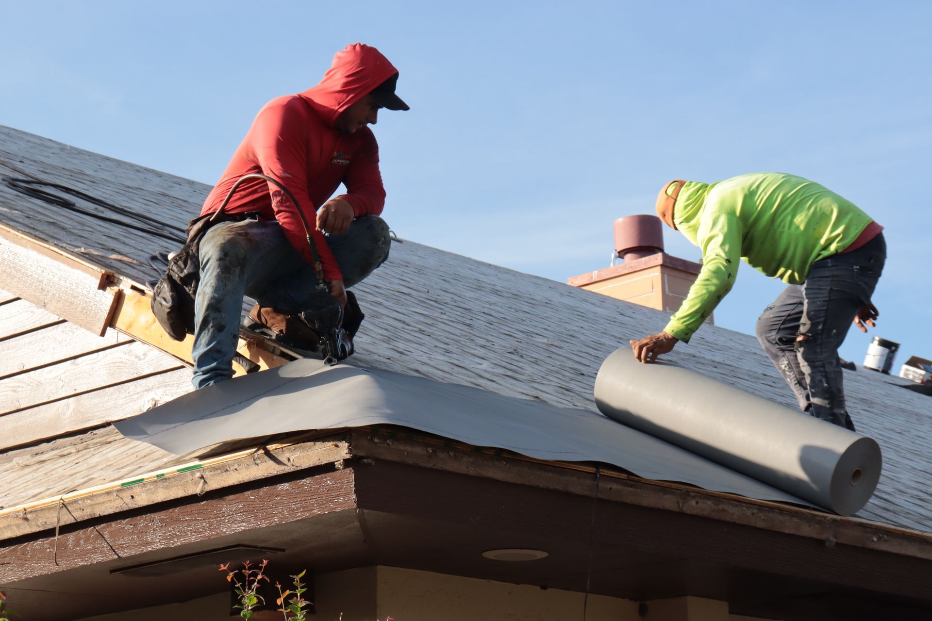 A person in a blue shirt applies sealant around a plumbing vent pipe on a flat, dark roof with many circular fasteners.