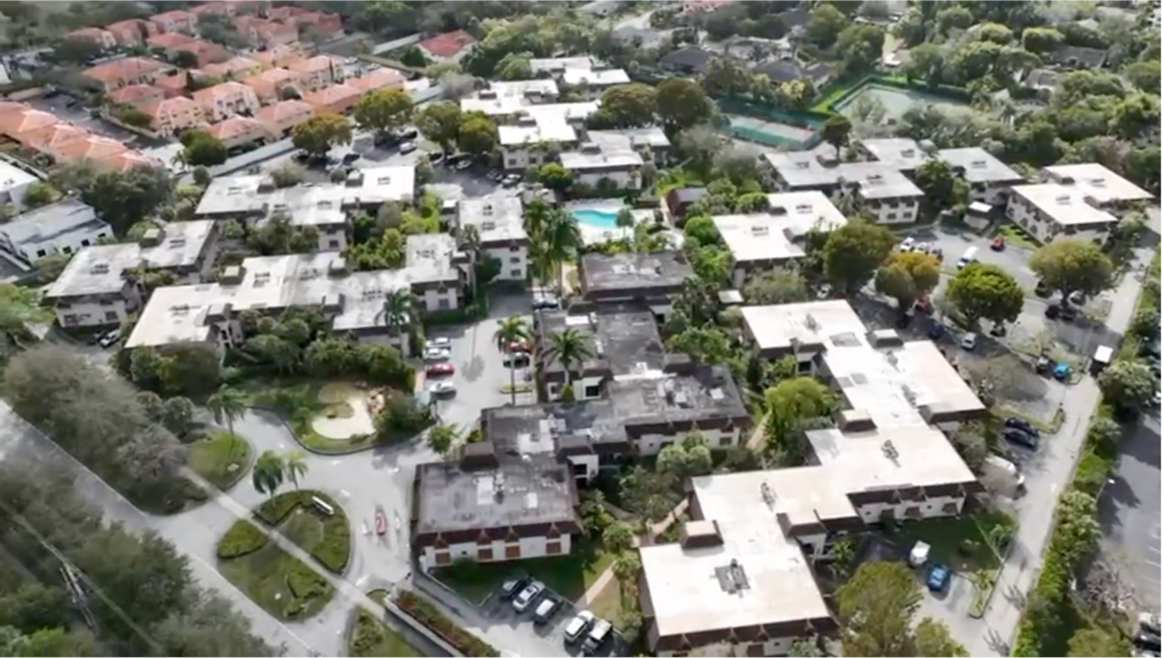 Four roofers in red long-sleeved shirts carry stacks of tiles across a sloped residential roof under a bright blue sky.