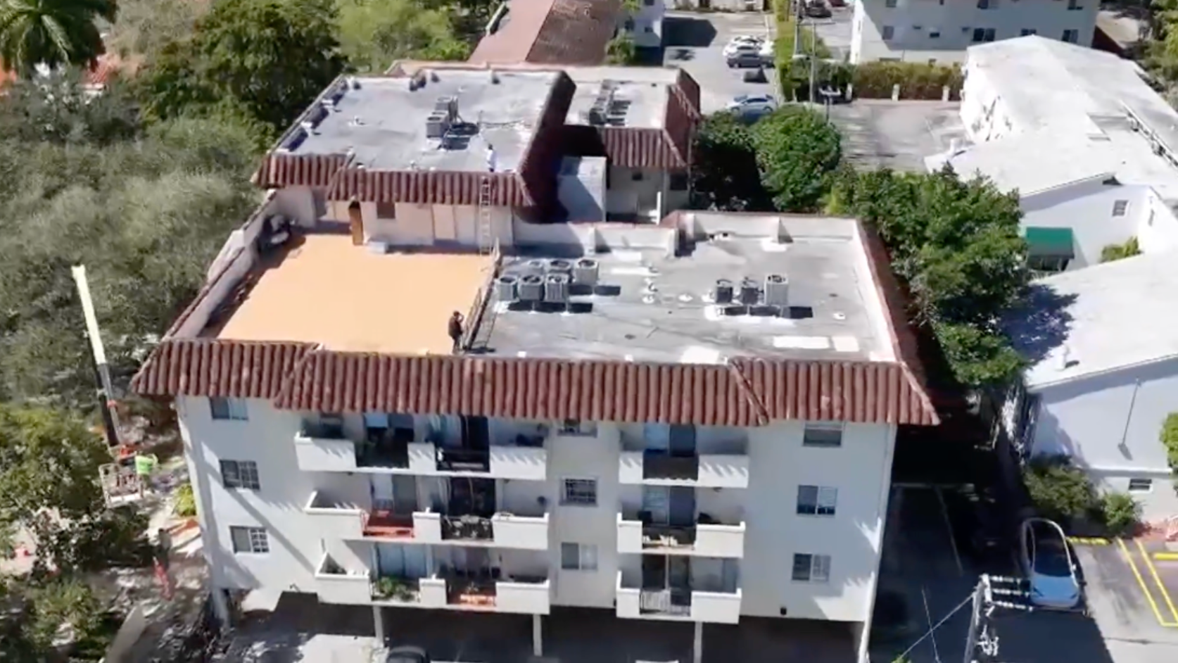 A person in a red shirt uses a pressure washer to clean dark, weathered clay roof tiles on a bright, sunny day.