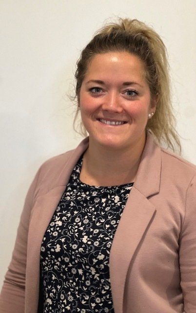 Une personne souriante, les cheveux blonds tirés en arrière, portant un haut à fleurs sous un blazer rose poudré, sur fond blanc.