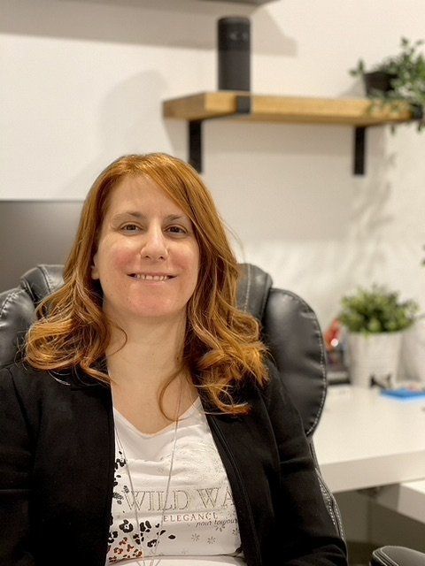 Une personne aux cheveux auburn mi-longs est assise sur une chaise de bureau, vêtue d'un blazer noir sur un t-shirt blanc à motifs.