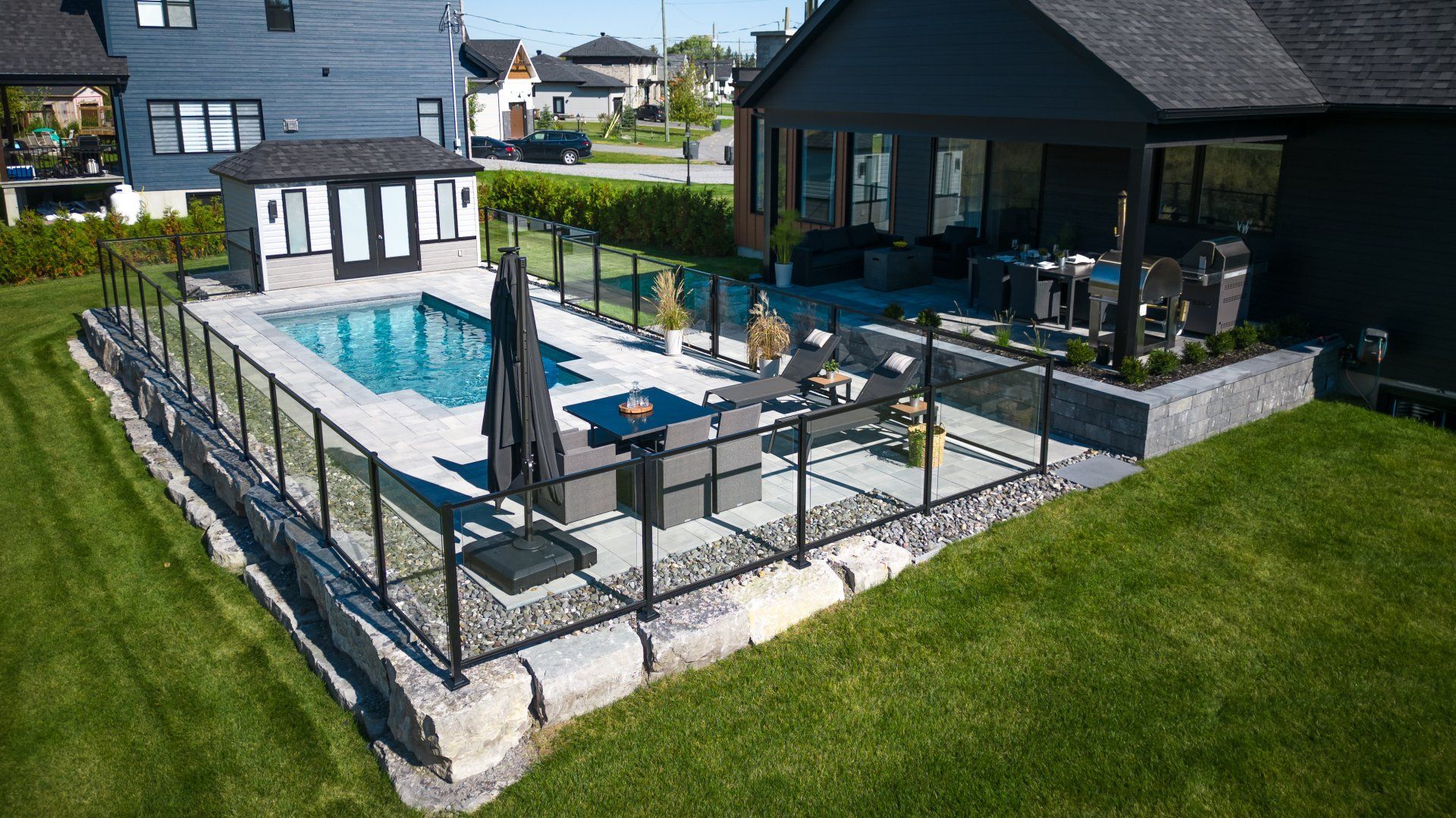 Vue aérienne d'un jardin comprenant une piscine, une terrasse entourée d'une clôture en verre, du mobilier de jardin et un cabanon.