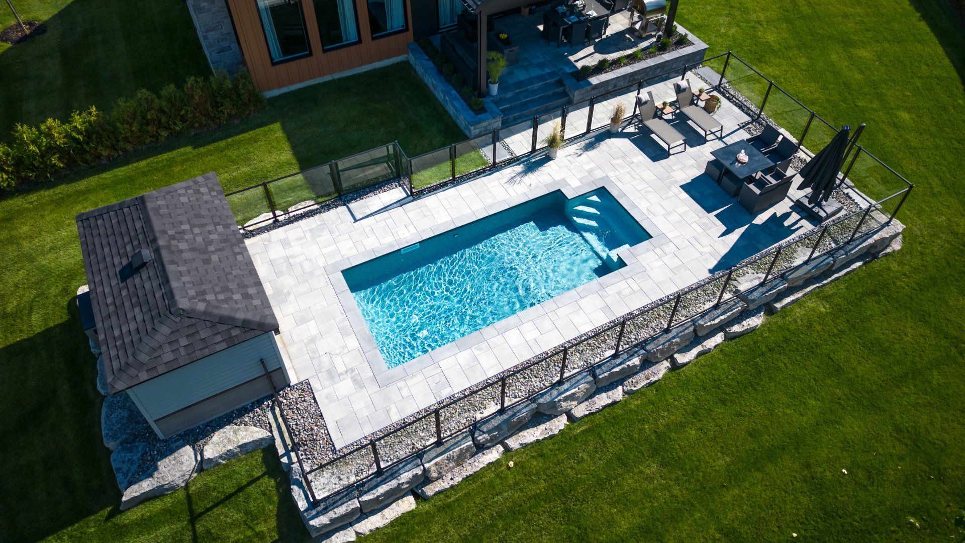 Vue aérienne d'une piscine rectangulaire dans un jardin avec une terrasse en pierre grise, un petit abri de jardin, des chaises longues et une clôture en verre.