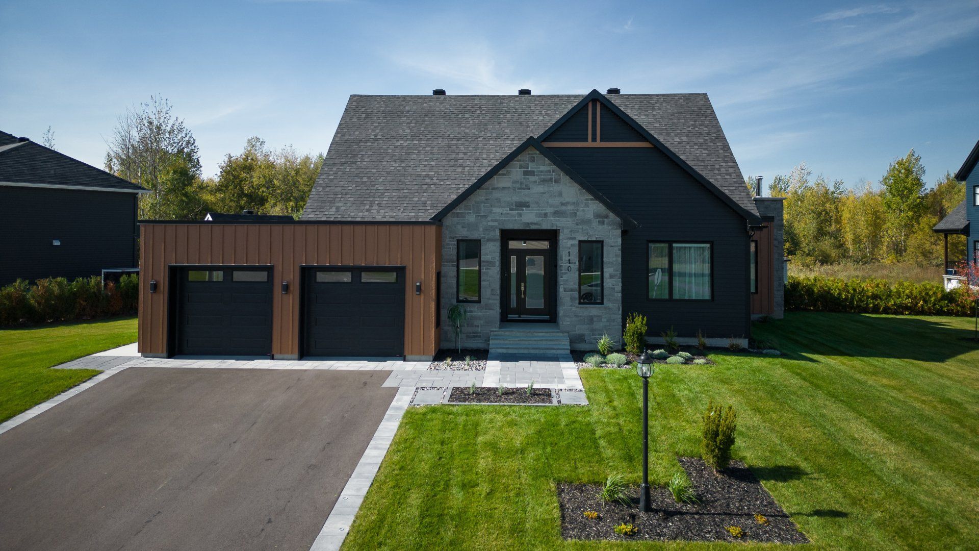 Une maison de banlieue moderne avec une façade en pierre grise, un bardage foncé, des éléments en bois dans le garage et une allée pavée, par une journée ensoleillée.