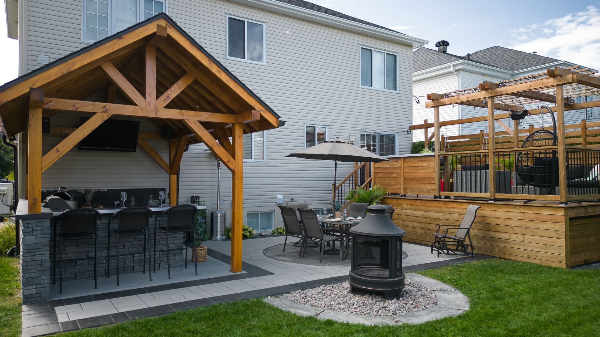 Une cour arrière de banlieue comprenant une cuisine extérieure couverte, une terrasse en pierre avec un ensemble de salle à manger, un foyer et une terrasse en bois surélevée.