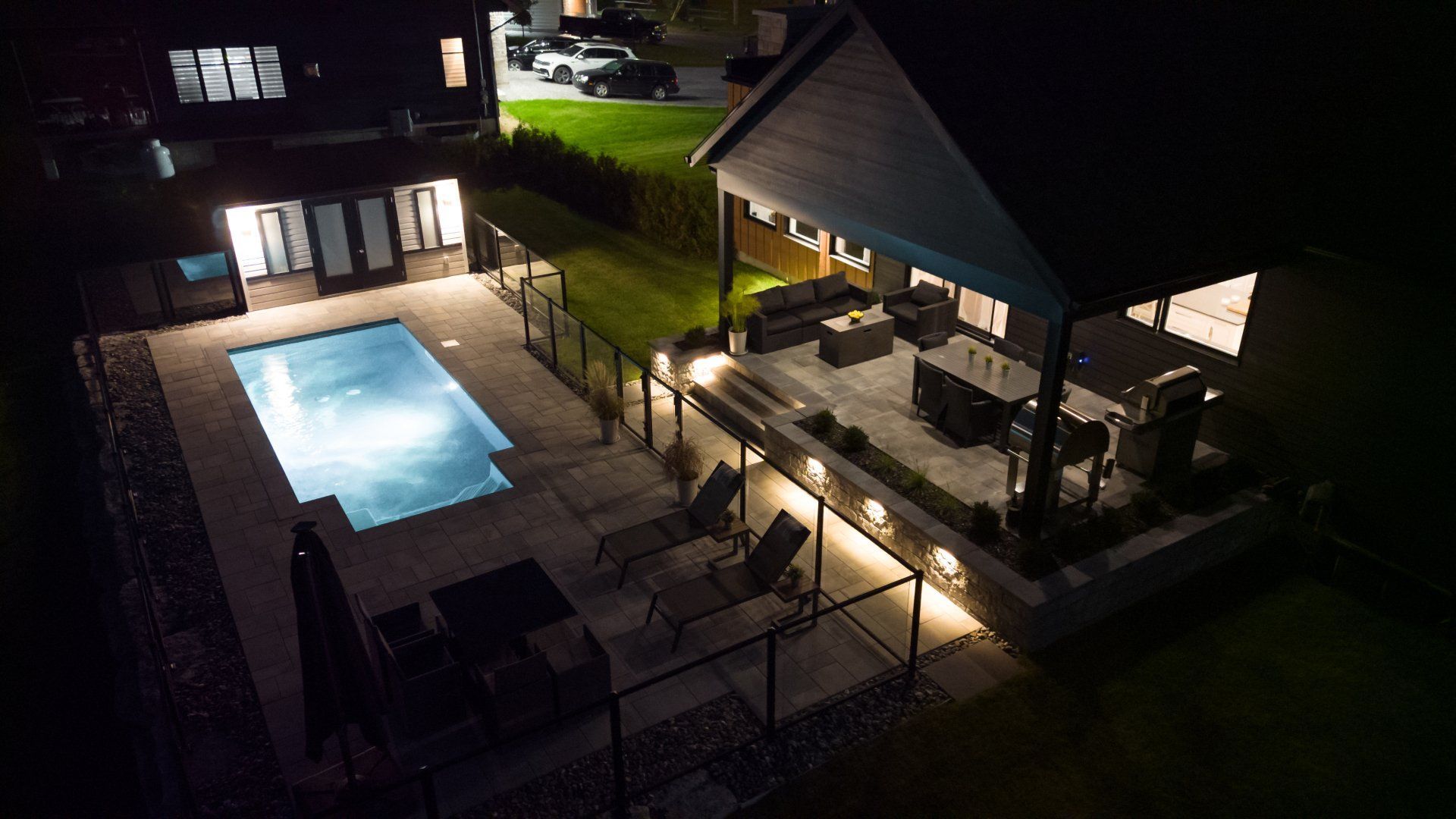 Vue aérienne nocturne d'une piscine illuminée dans un jardin, d'une terrasse en pierre et d'un coin salon extérieur couvert avec mobilier.