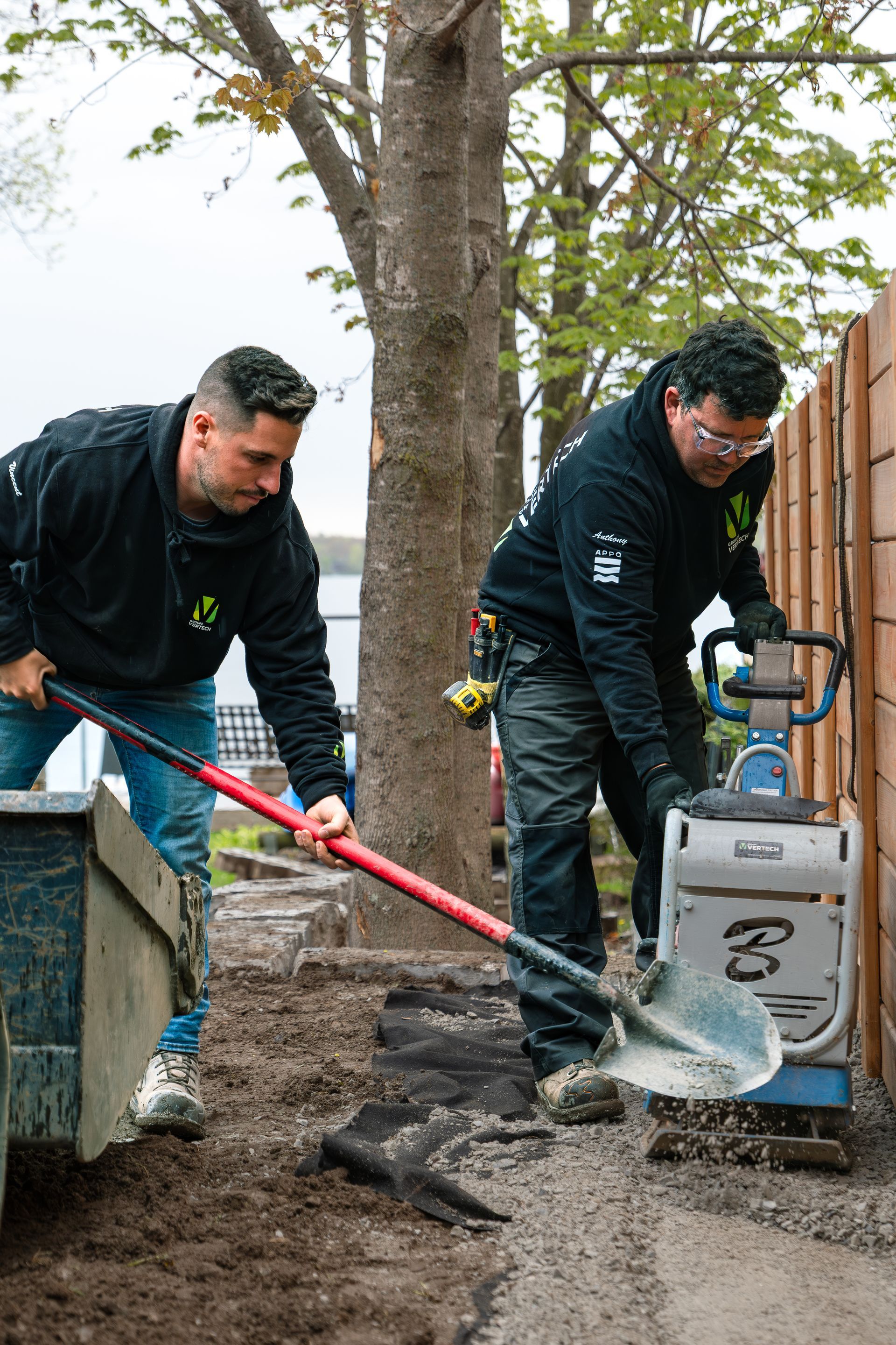Une piscine rectangulaire dans le jardin, entourée de dalles de pierre gris clair, d'une clôture en verre et d'une terrasse ombragée.