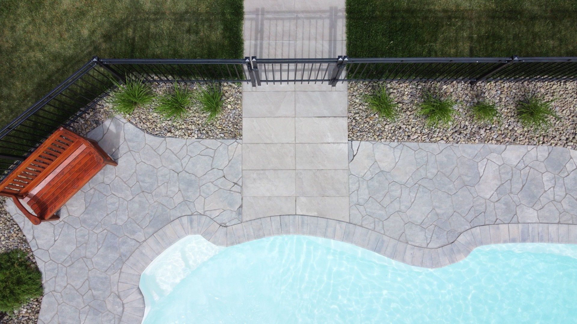 Vue aérienne d'une piscine dans un jardin, d'une terrasse en pierre, d'un banc en bois, d'une allée centrale et d'une clôture noire.