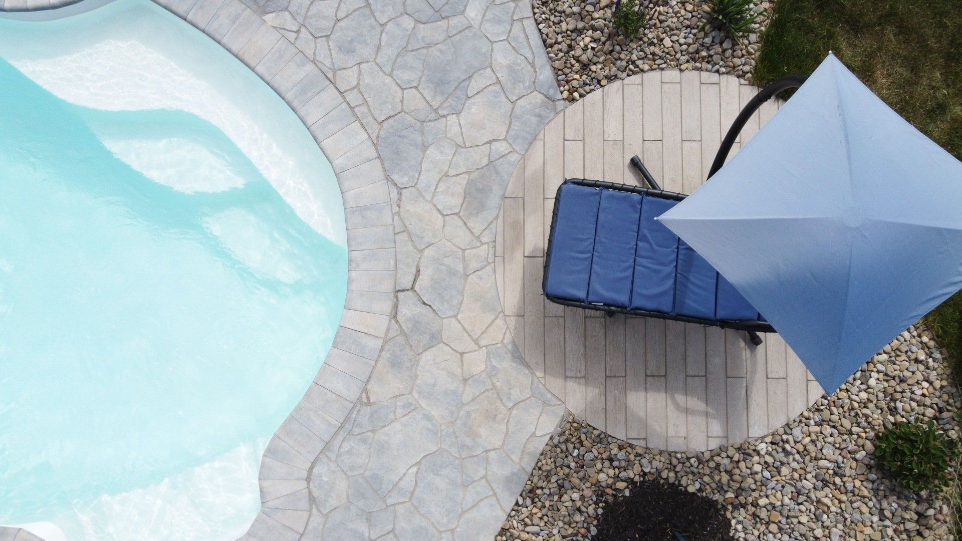 Vue en plongée d'une piscine à l'eau bleu clair, d'une terrasse en pierre et d'une chaise longue bleue sous un parasol.