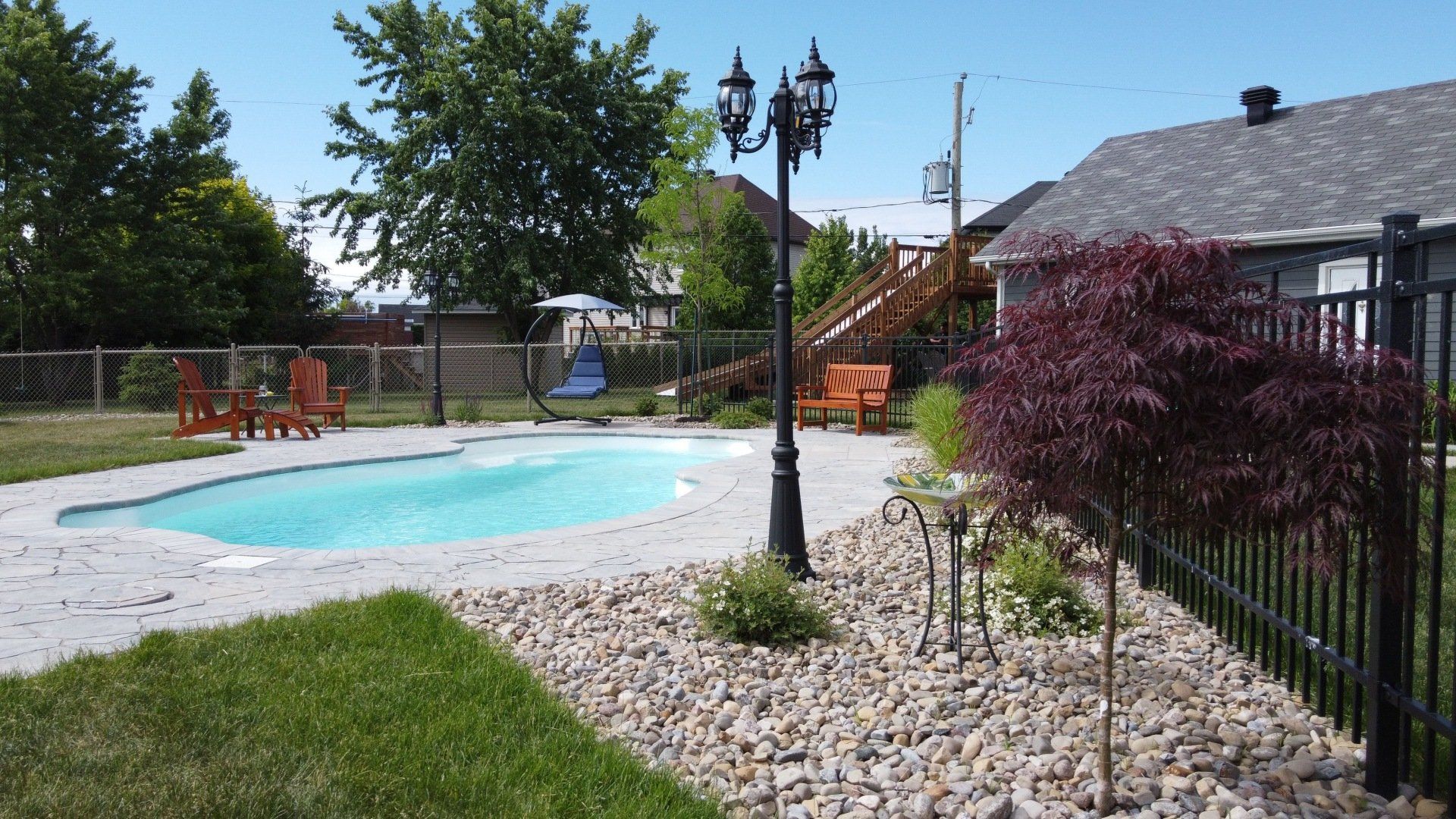 Une piscine dans un jardin avec une terrasse en béton, un aménagement paysager en pierre, un lampadaire noir et une clôture, par une journée ensoleillée.
