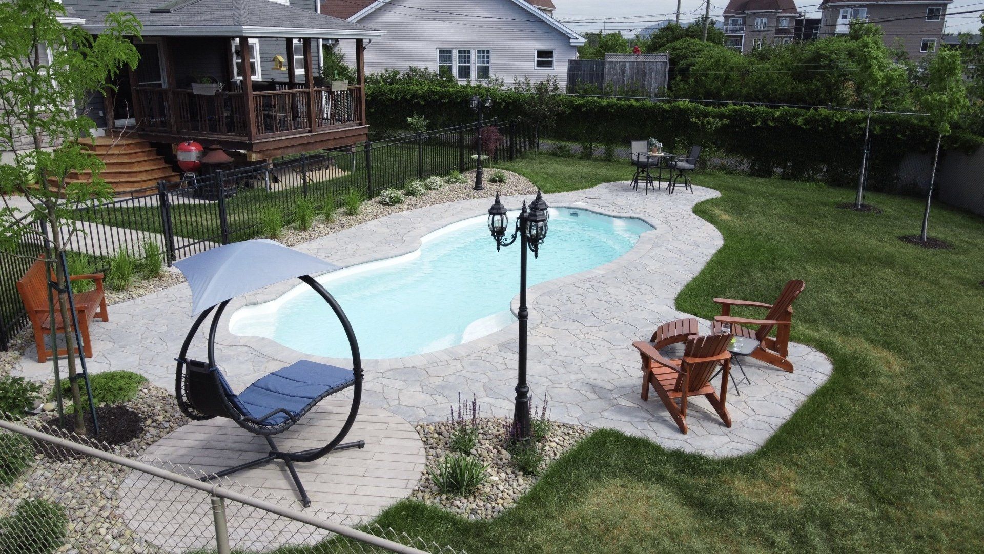 Un jardin avec une piscine en forme de haricot, une terrasse en pierre, du mobilier de jardin et un lampadaire sous un ciel nuageux.