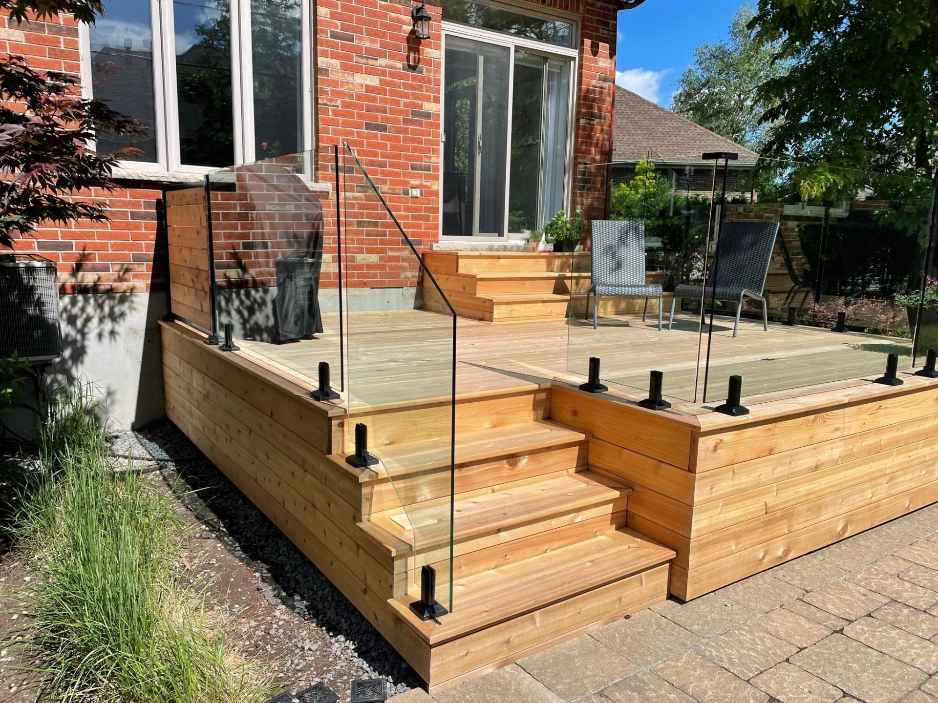 Une terrasse en bois avec une rambarde en verre et des marches menant à une maison en briques avec une porte coulissante en verre et du mobilier de jardin.