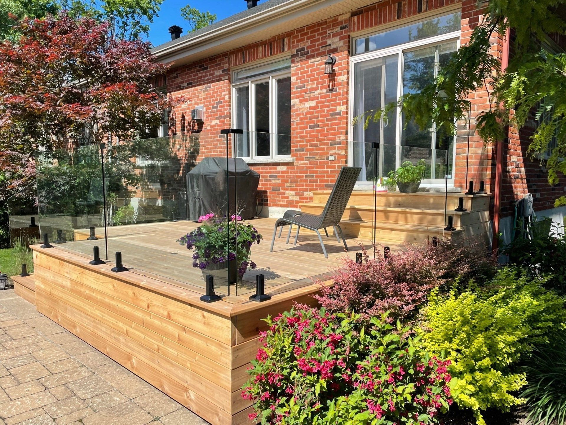 Une terrasse en bois avec une rambarde en verre, des meubles de jardin et des plantes en pot devant une maison en briques rouges par une journée ensoleillée.