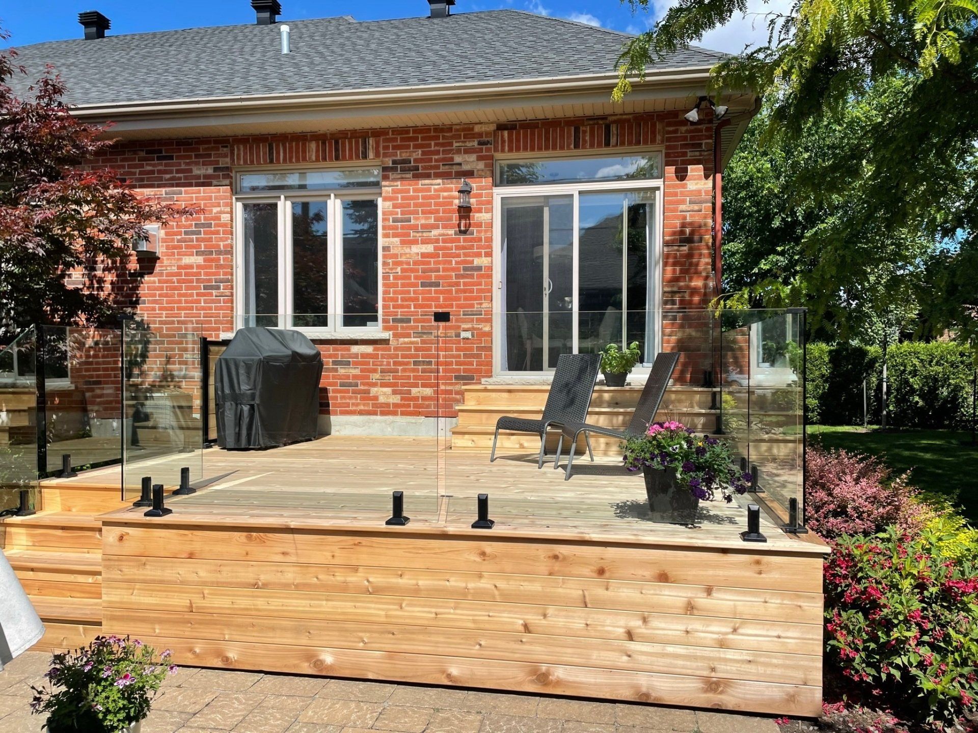 Une terrasse en bois avec une balustrade en verre et deux chaises longues se trouve à l'extérieur d'une maison en briques dotée d'une porte-fenêtre coulissante.