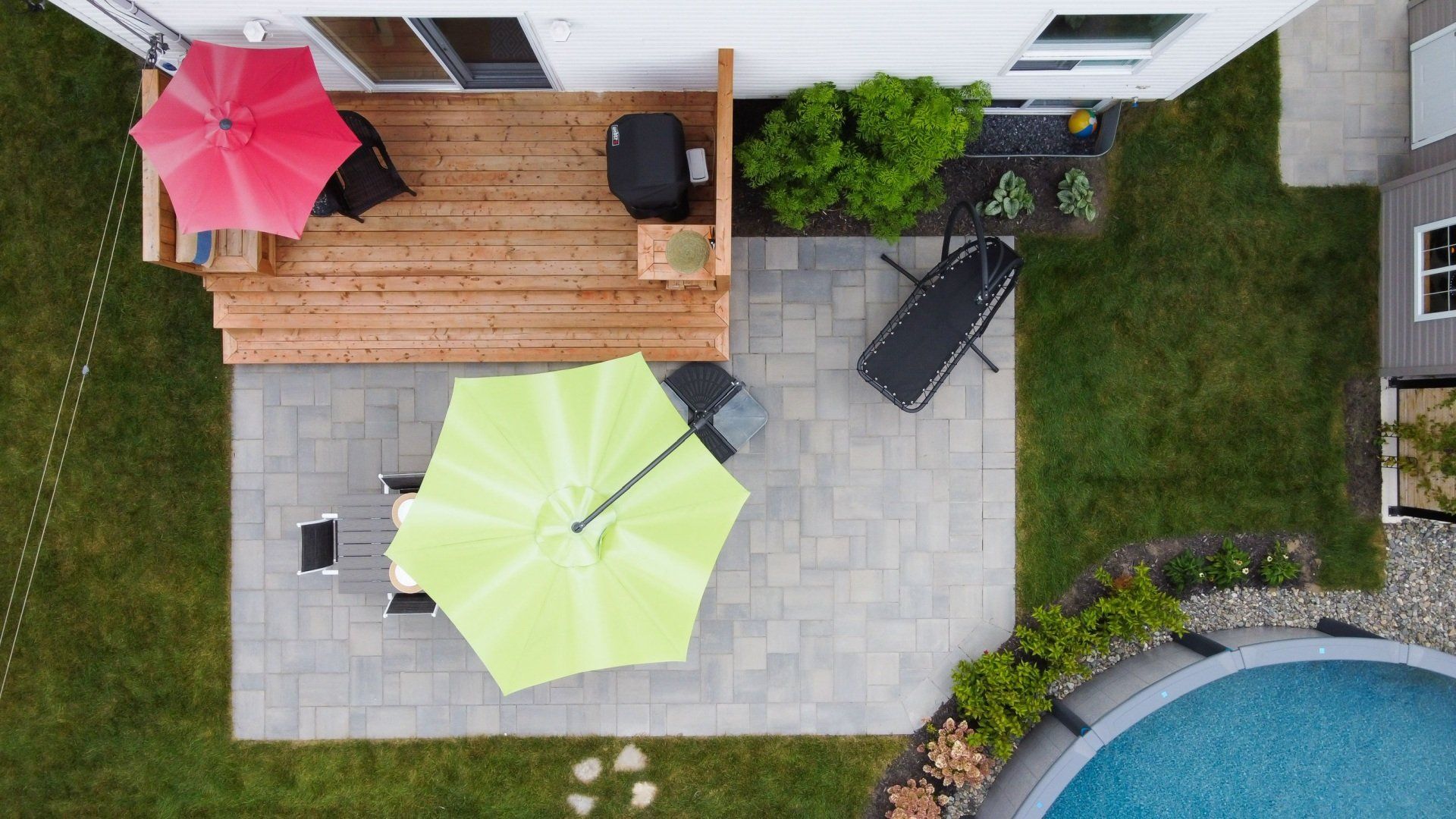 Vue aérienne d'une terrasse en bois avec un parasol rouge et d'un patio en pierre avec un parasol vert clair à côté d'une piscine.