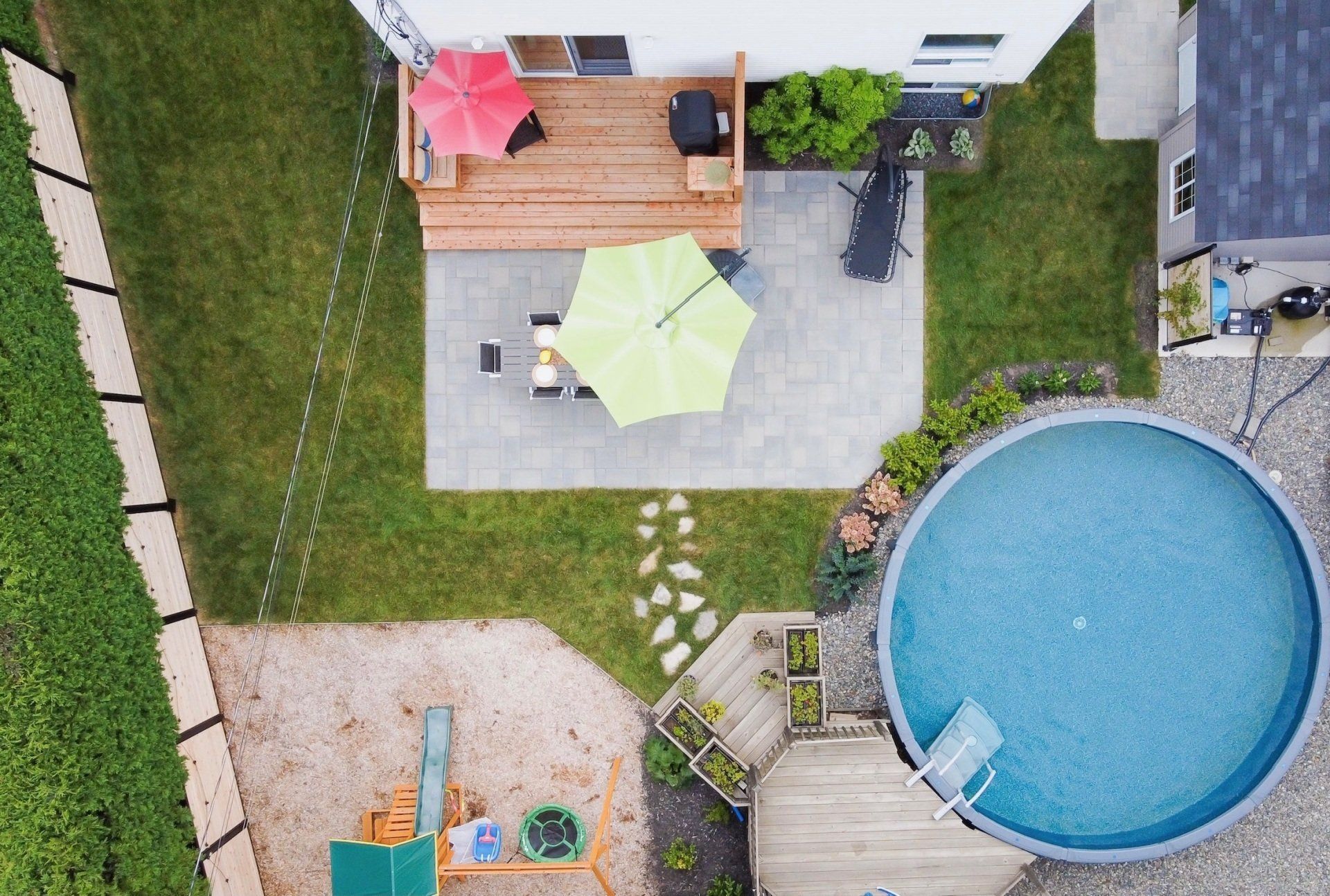 Vue aérienne d'une cour arrière avec une terrasse, un patio en pierre, une pelouse verte, une piscine hors sol et une aire de jeux en gravier.