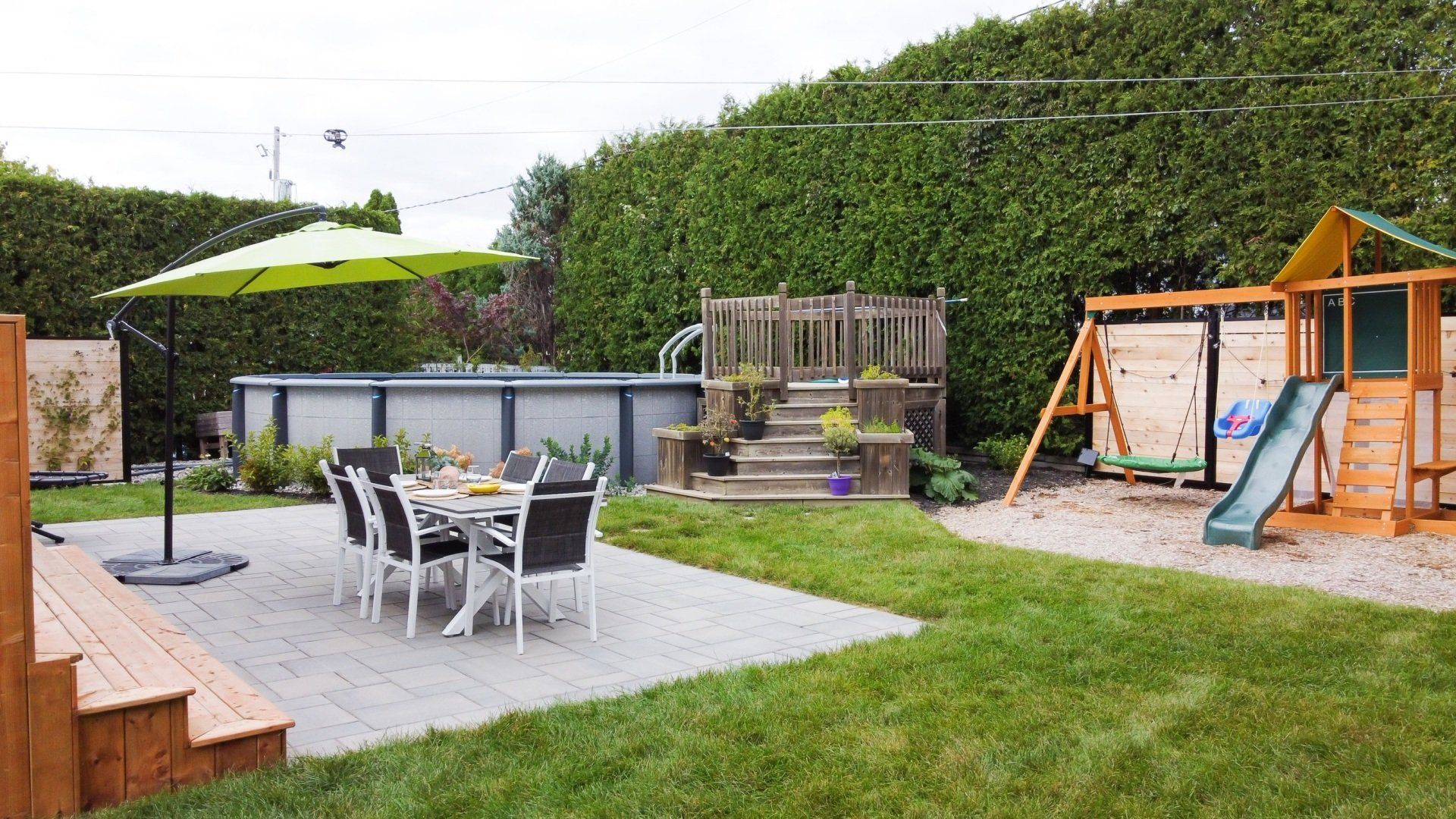 Un jardin avec une terrasse ombragée par un parasol vert citron, une piscine hors sol circulaire et une balançoire en bois sur du gravier.