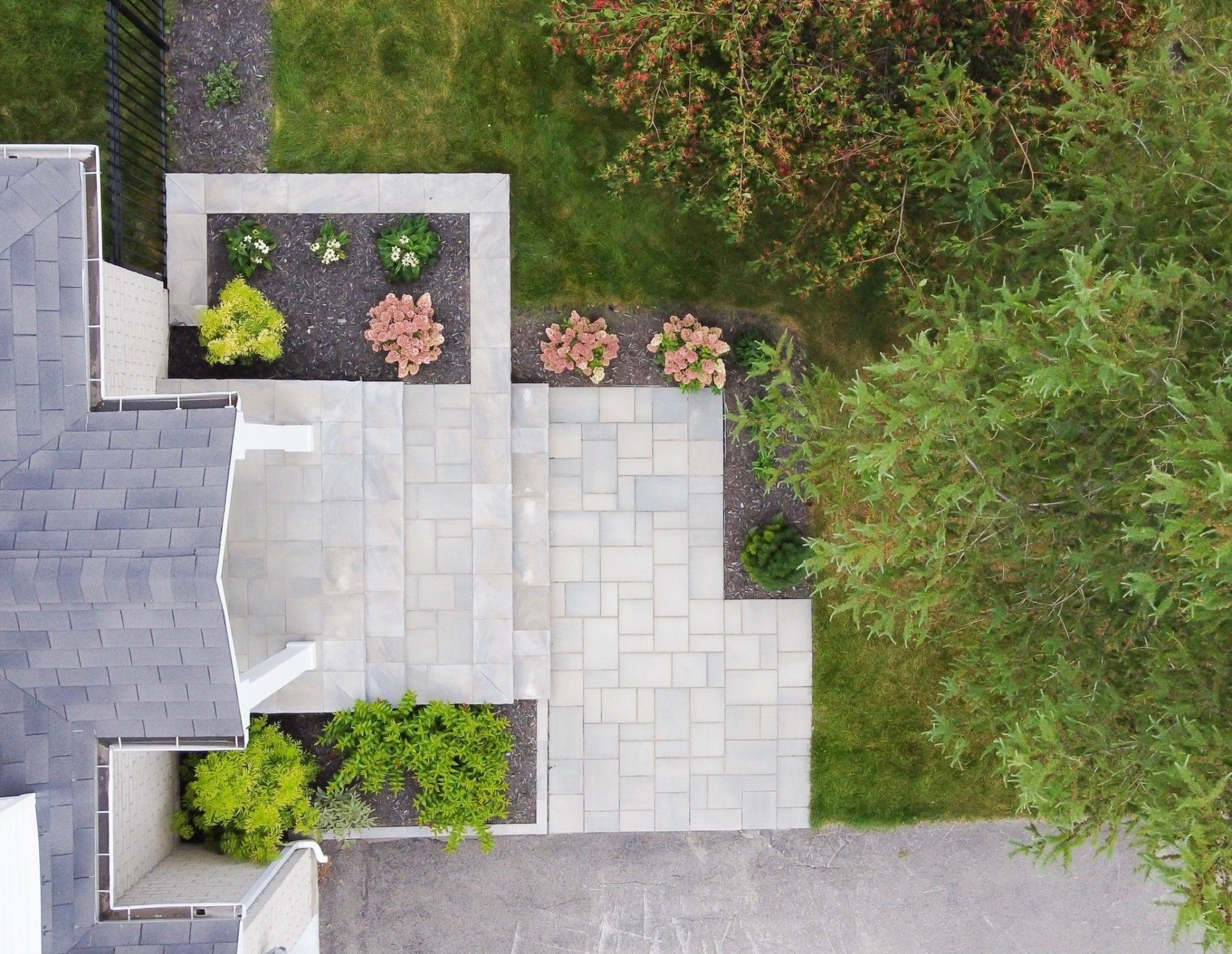 Vue aérienne d'une terrasse et d'une allée pavées de couleur claire, bordées de parterres de fleurs avec des plantes et des arbustes, à côté d'une maison.