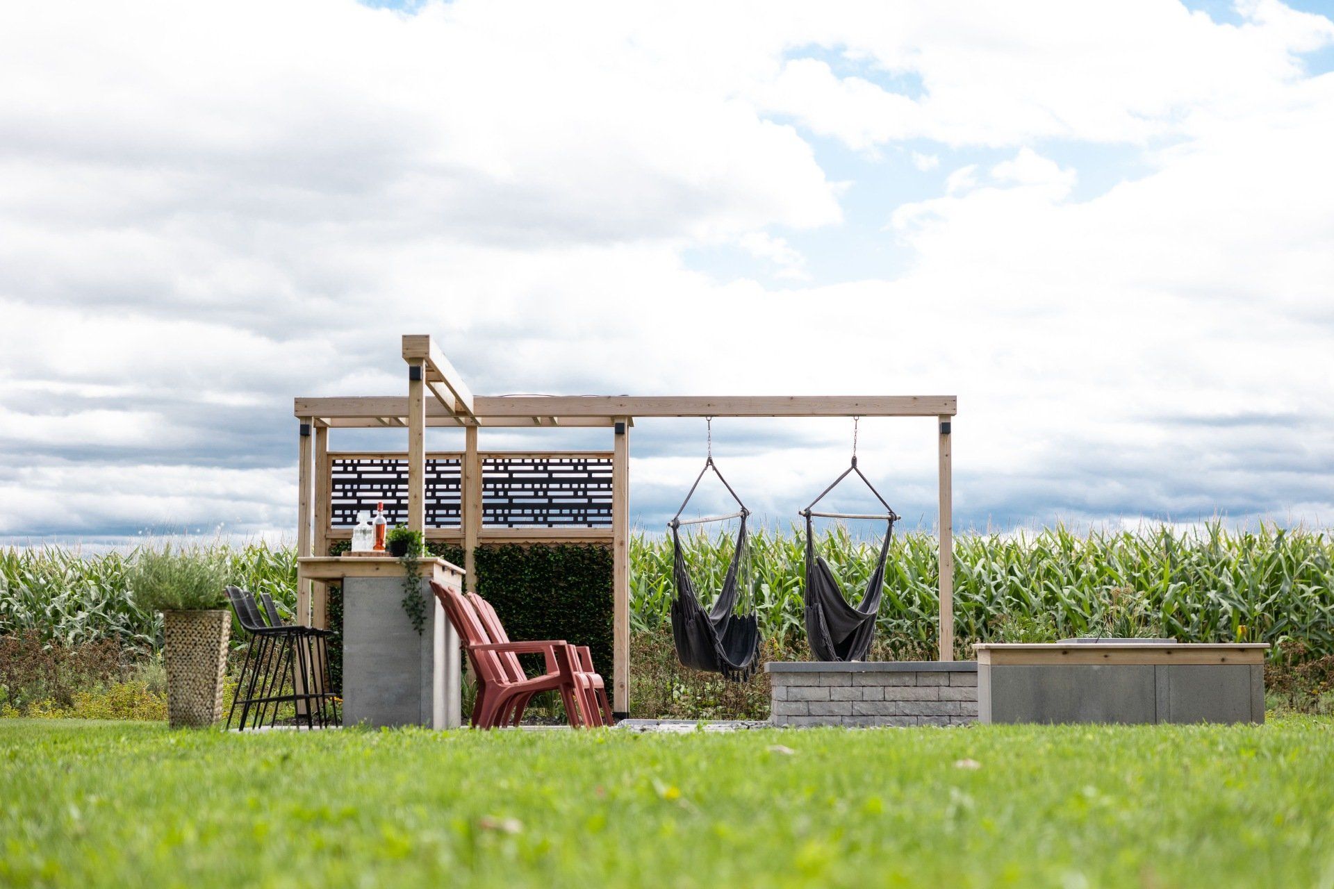 Un salon de jardin en bois avec un petit bar, des chaises rouges et deux chaises suspendues, situé devant un champ de maïs.