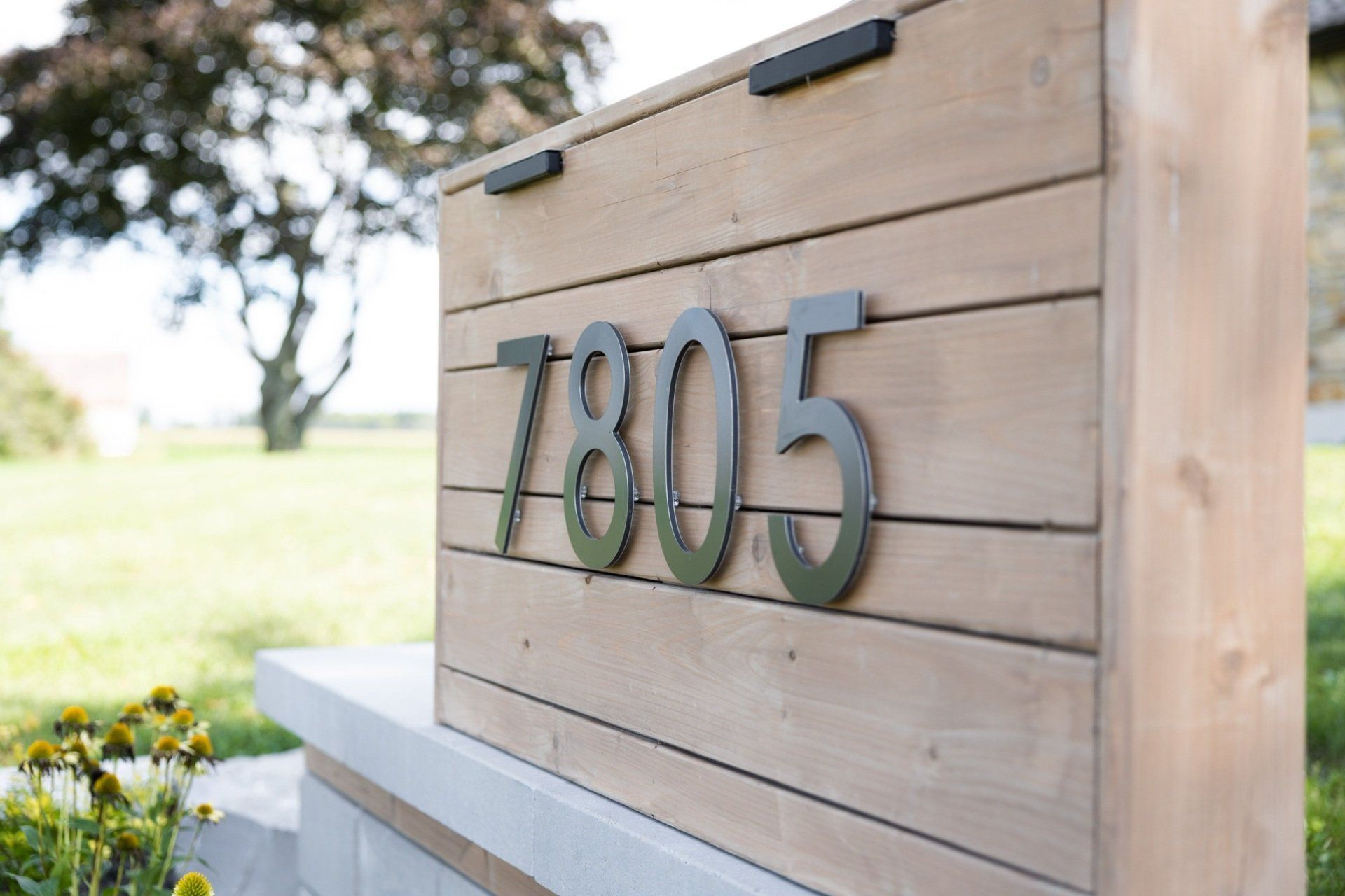 Une plaque de maison moderne en bois affichant le numéro d'adresse 7805 en métal foncé, sur un fond herbeux flou.