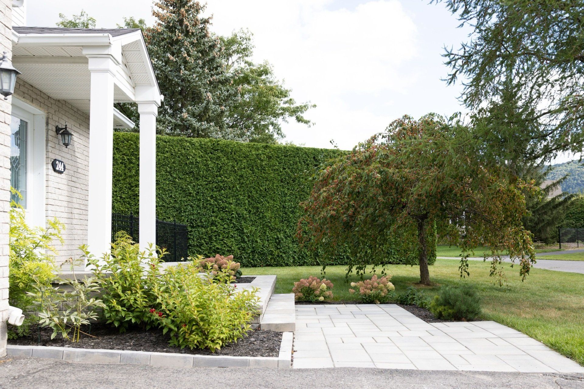 L'entrée de cette maison en briques claires est ornée de colonnes blanches, d'une terrasse pavée, d'aménagements paysagers et d'une haute haie verte assurant l'intimité.