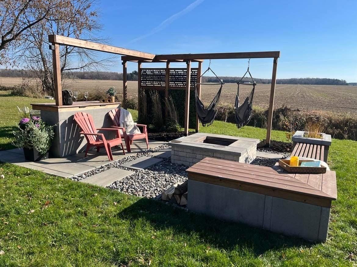 Terrasse extérieure avec foyer, chaises rouges, sièges suspendus et bar en béton, donnant sur un vaste champ.