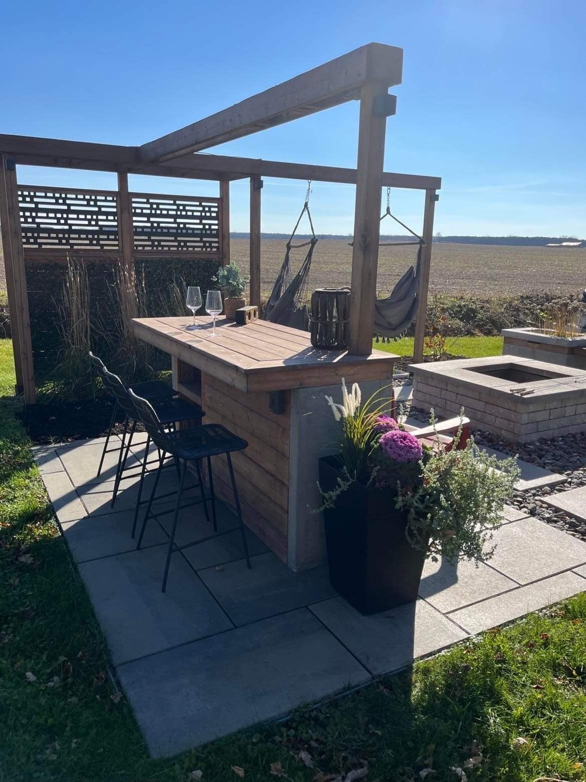 Un patio extérieur avec une pergola en bois, des tabourets de bar, un foyer et des chaises suspendues donnant sur un champ.