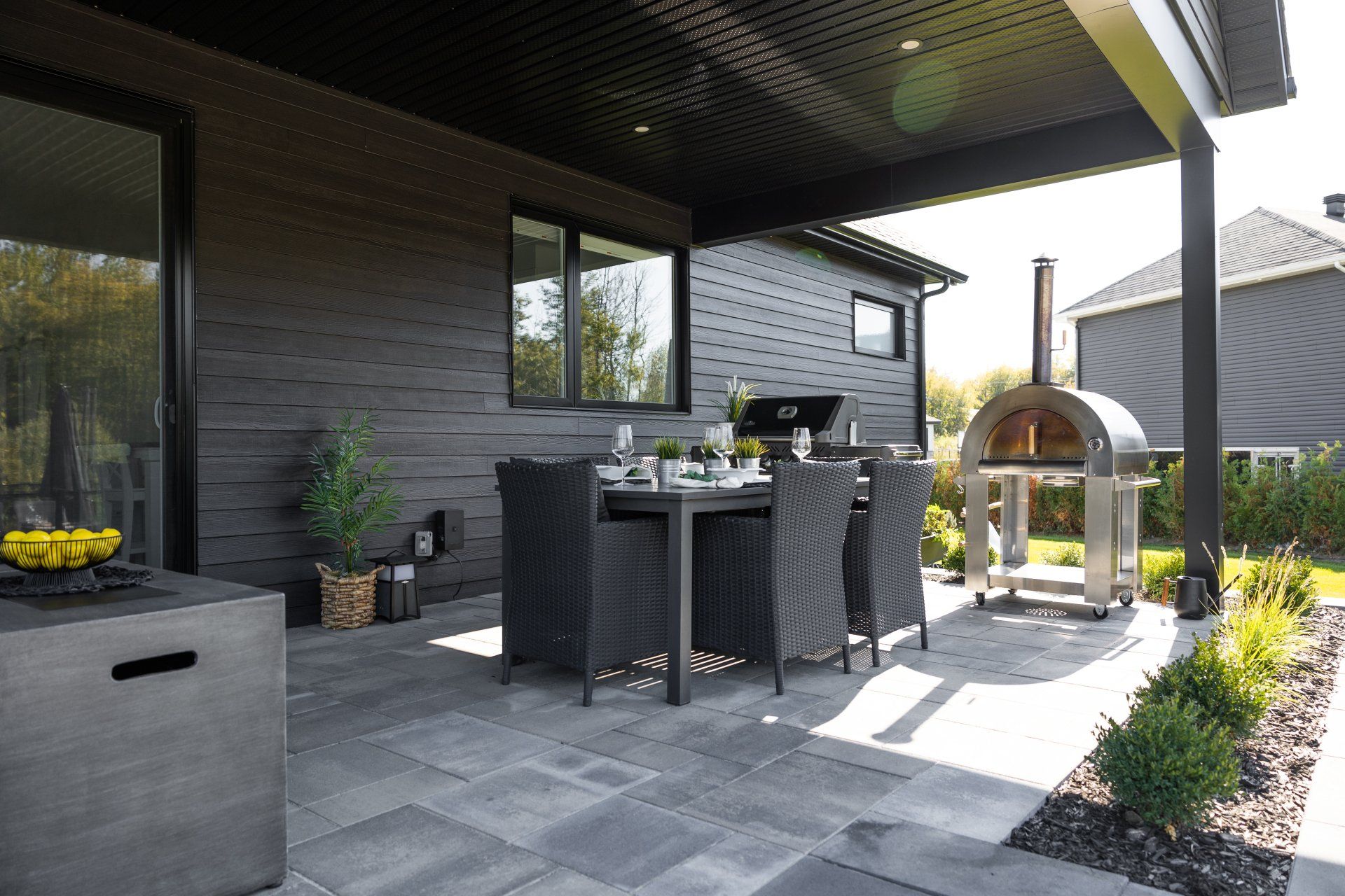 Une terrasse extérieure moderne comprenant une table à manger avec des chaises, un four extérieur et un revêtement extérieur foncé sous un toit couvert.