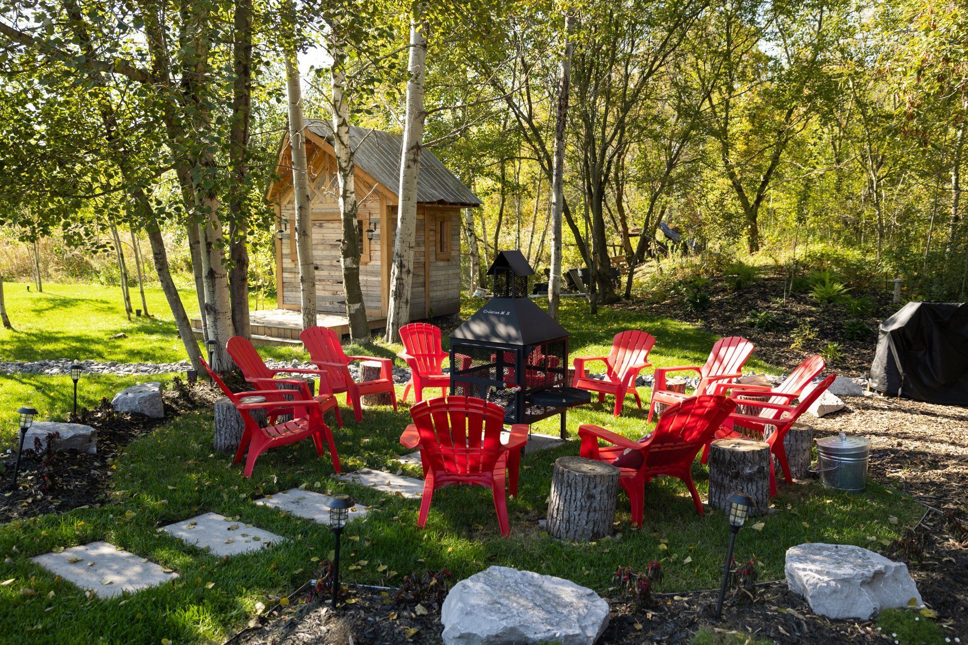 Un cercle de chaises Adirondack rouge vif entoure un foyer en métal noir dans un espace herbeux avec une petite cabane en bois.