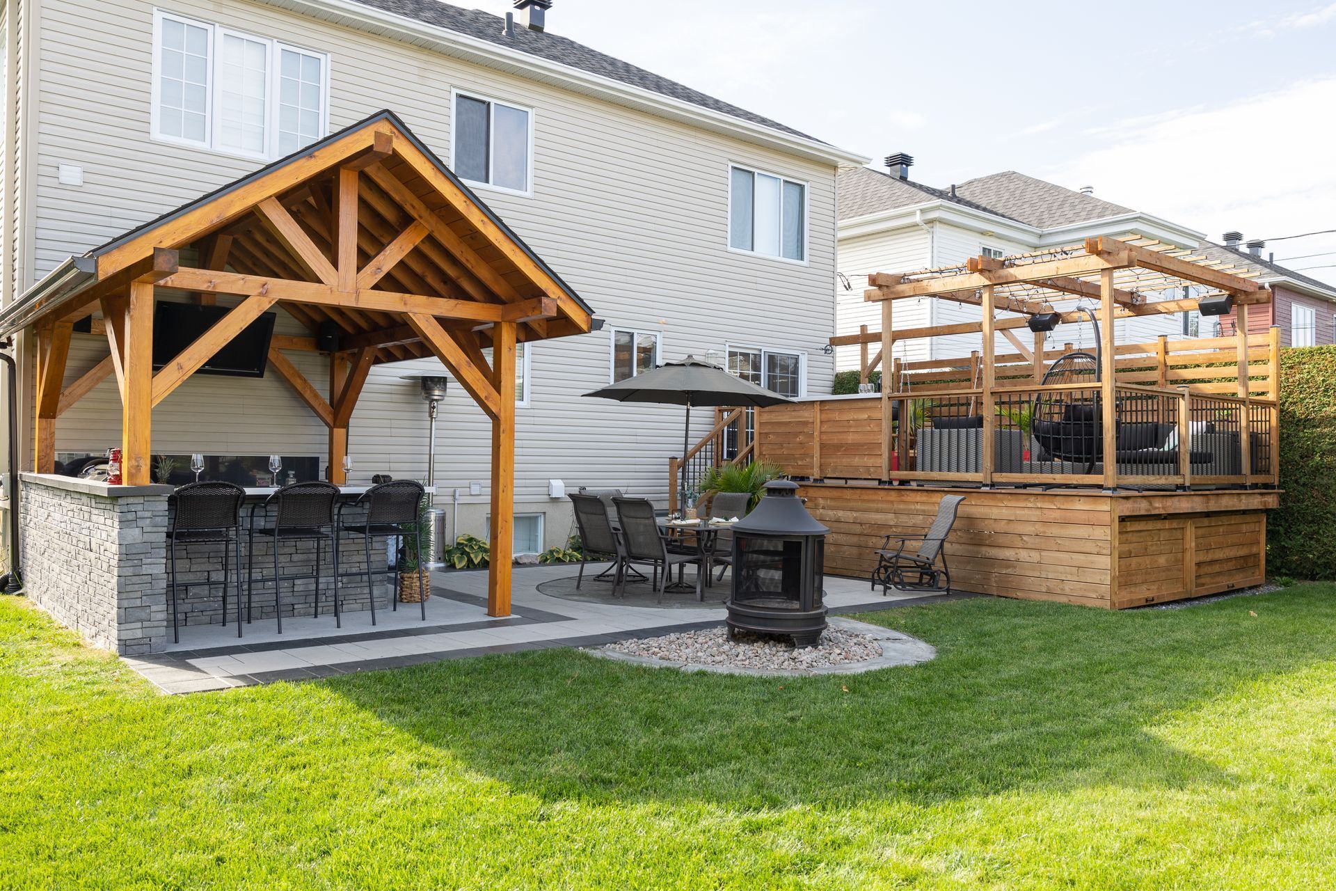 Terrasse arrière avec cuisine extérieure à ossature bois, base en pierre, ensemble de table et chaises, foyer et terrasse en bois surélevée avec pergola.