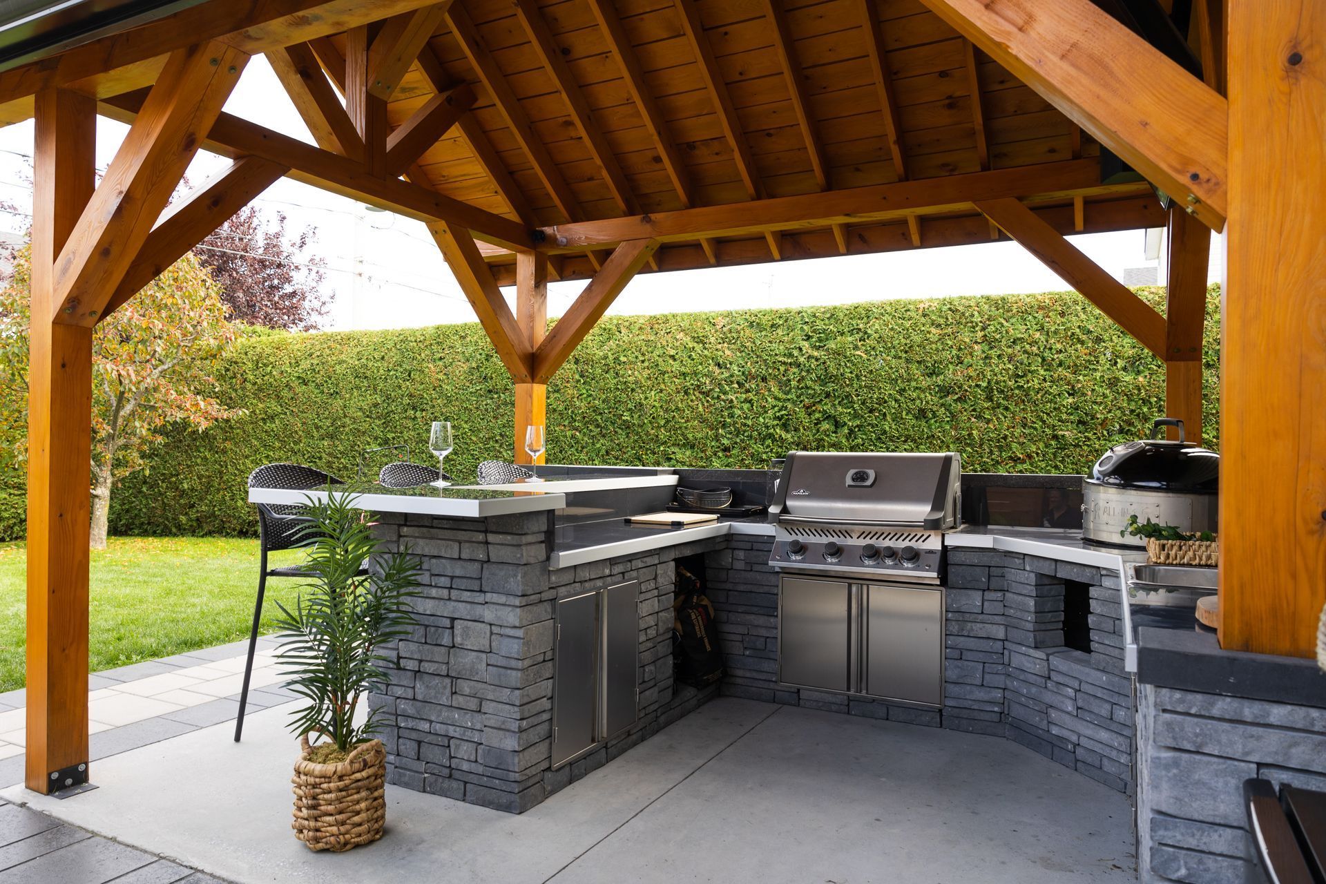 Une cuisine extérieure avec un îlot revêtu de pierre, un barbecue en acier inoxydable et des sièges de bar sous un gazebo en bois sur une terrasse.