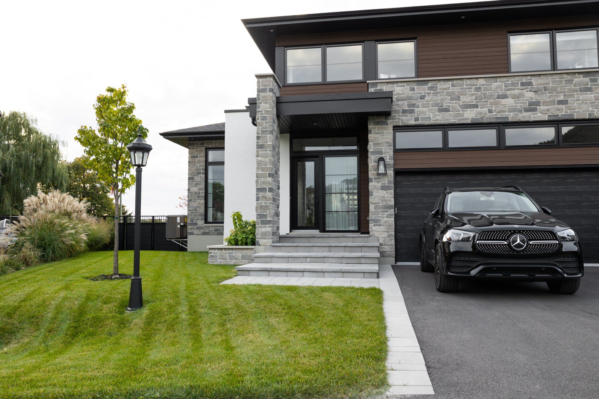 Maison moderne de deux étages en pierre et bois foncé, avec une Mercedes noire garée dans l'allée et un lampadaire sur une pelouse verte.