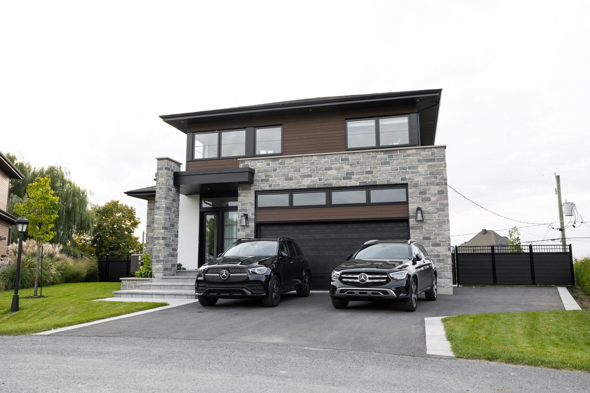 Une maison moderne à deux étages avec un revêtement en pierre et un toit foncé, comprenant un garage double et des SUV noirs garés dans l'allée.