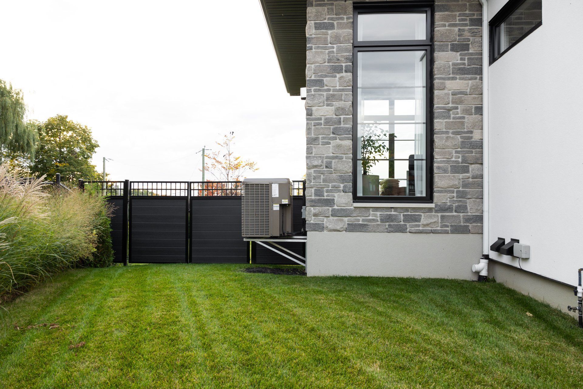 L'extérieur d'une maison en pierre, avec une clôture noire, une climatisation centrale et un jardin gazonné.