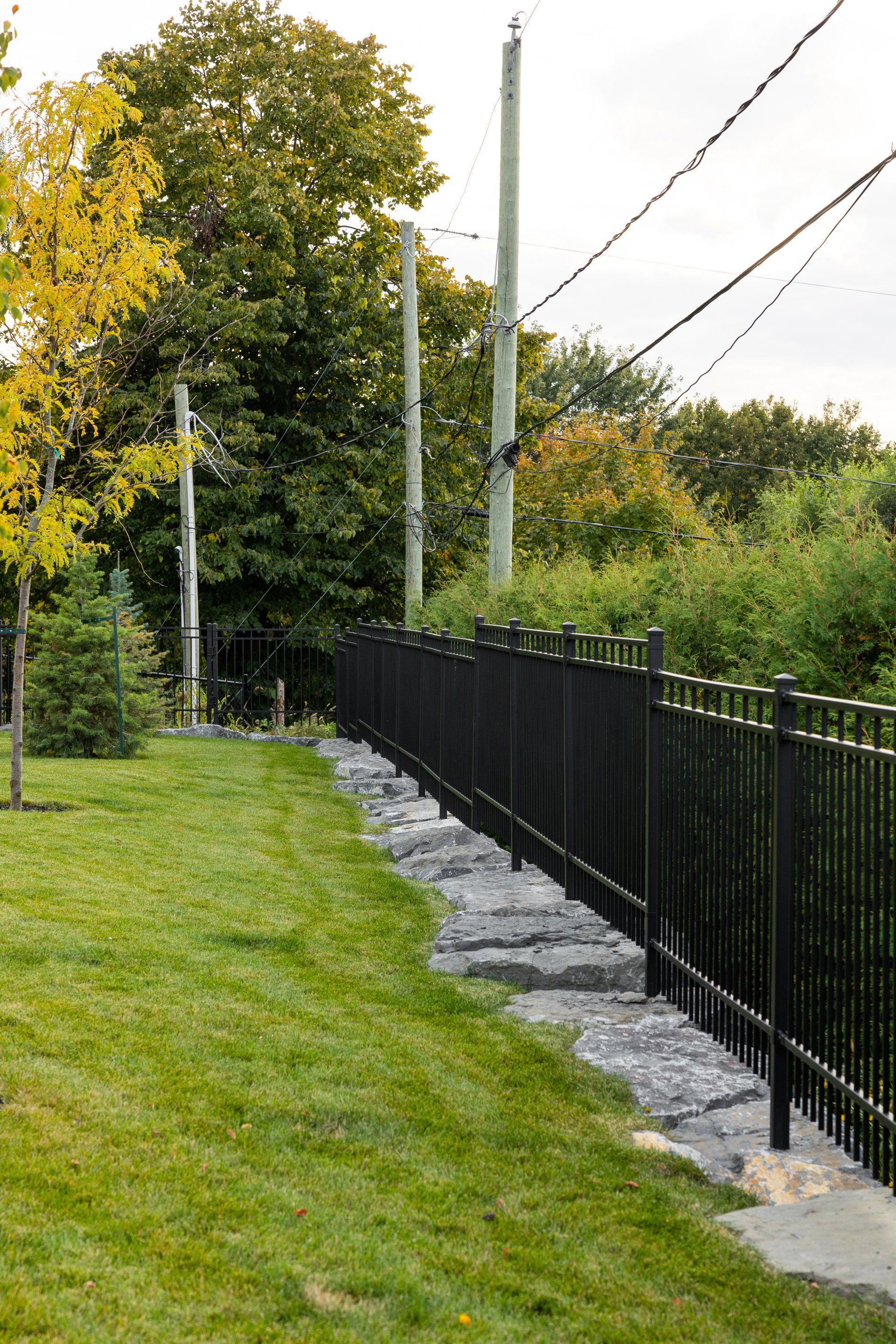 Une clôture en métal noir longe un socle en pierre dans une cour herbeuse, avec des poteaux électriques et des arbres en arrière-plan.