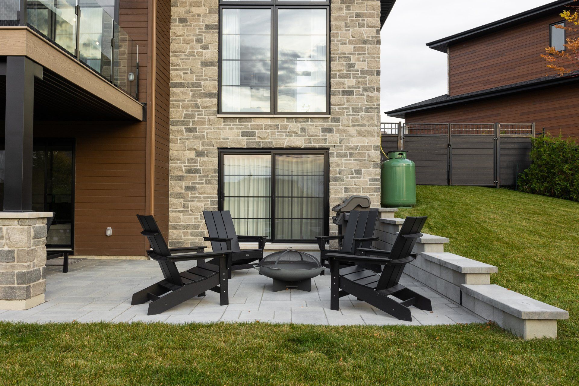 Terrasse avec quatre chaises Adirondack noires et un foyer, adossée à une maison en pierre et donnant sur un jardin gazonné.