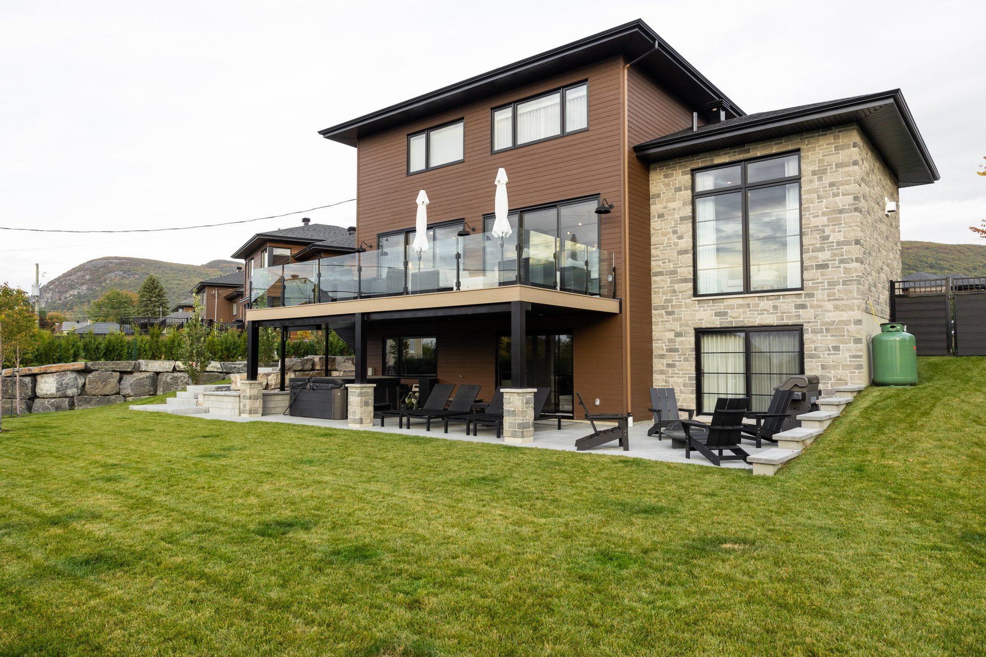 Une maison moderne de deux étages avec une façade en pierre, un bardage brun, une terrasse spacieuse avec des balustrades en verre et un jardin gazonné à l'arrière.