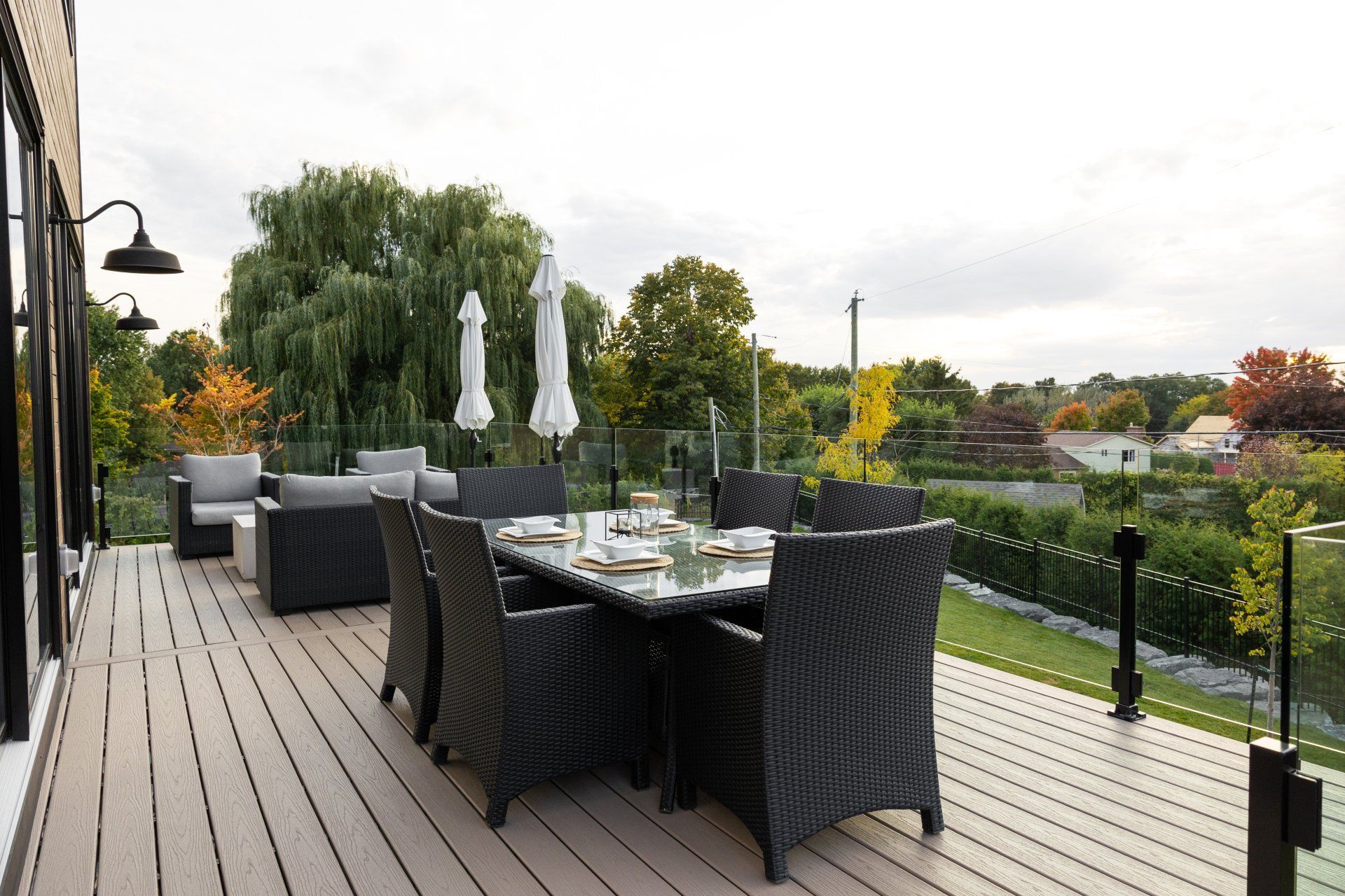 Par temps nuageux, une terrasse en bois avec un ensemble de salle à manger en osier sombre et des chaises longues surplombe un jardin luxuriant et arboré.