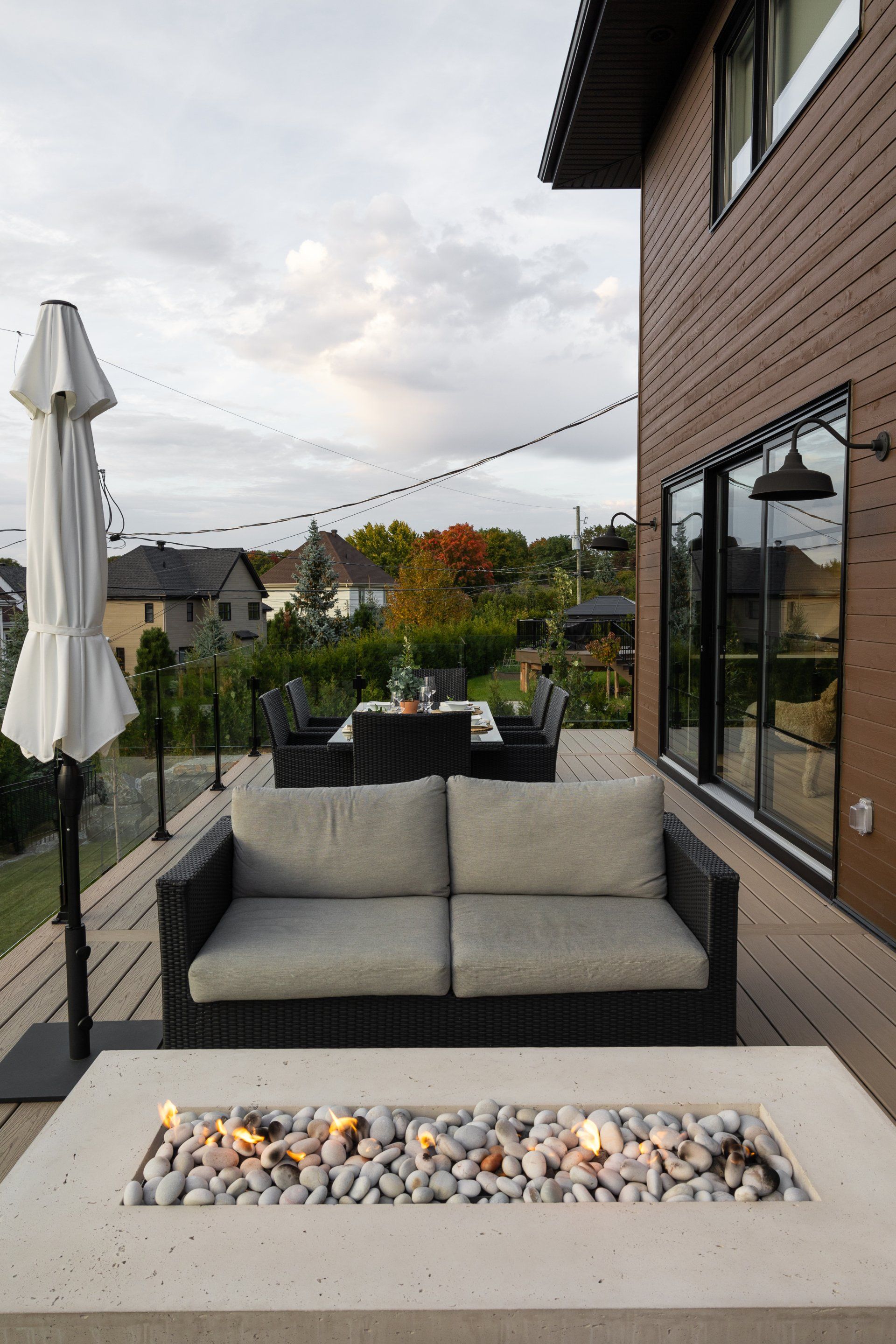Une terrasse moderne avec un foyer, un canapé confortable et un ensemble de repas en plein air, située à côté d'une maison sous un ciel nuageux.