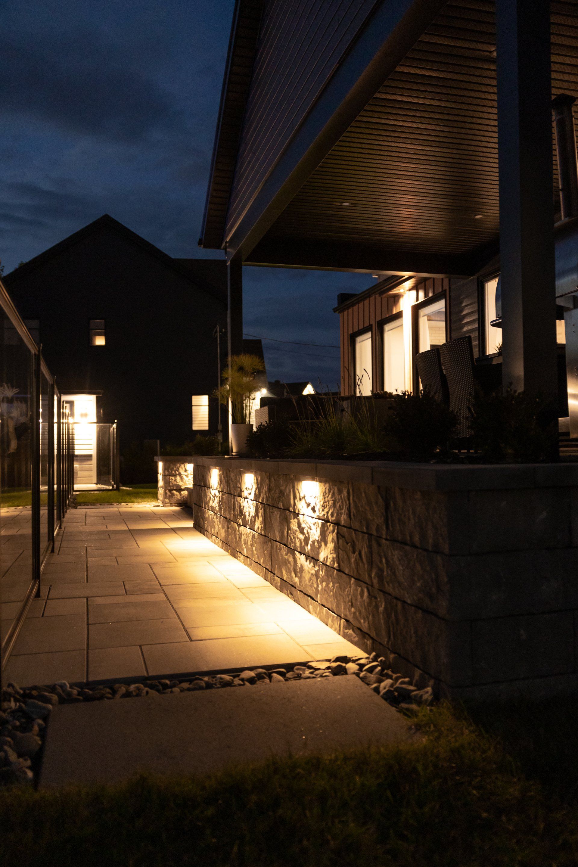 Une terrasse de jardin au crépuscule, illuminée par des lampes de jardin encastrées dans un mur de soutènement en pierre.