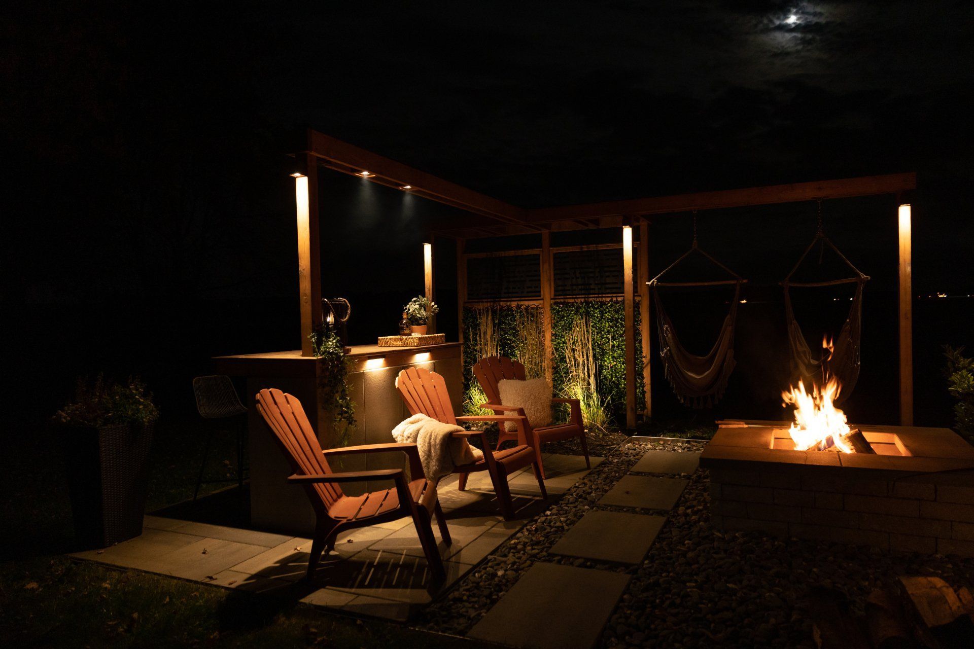 Une terrasse extérieure accueillante le soir, avec deux chaises en bois, un foyer, un comptoir de bar et des balançoires suspendues sous un éclairage chaleureux.